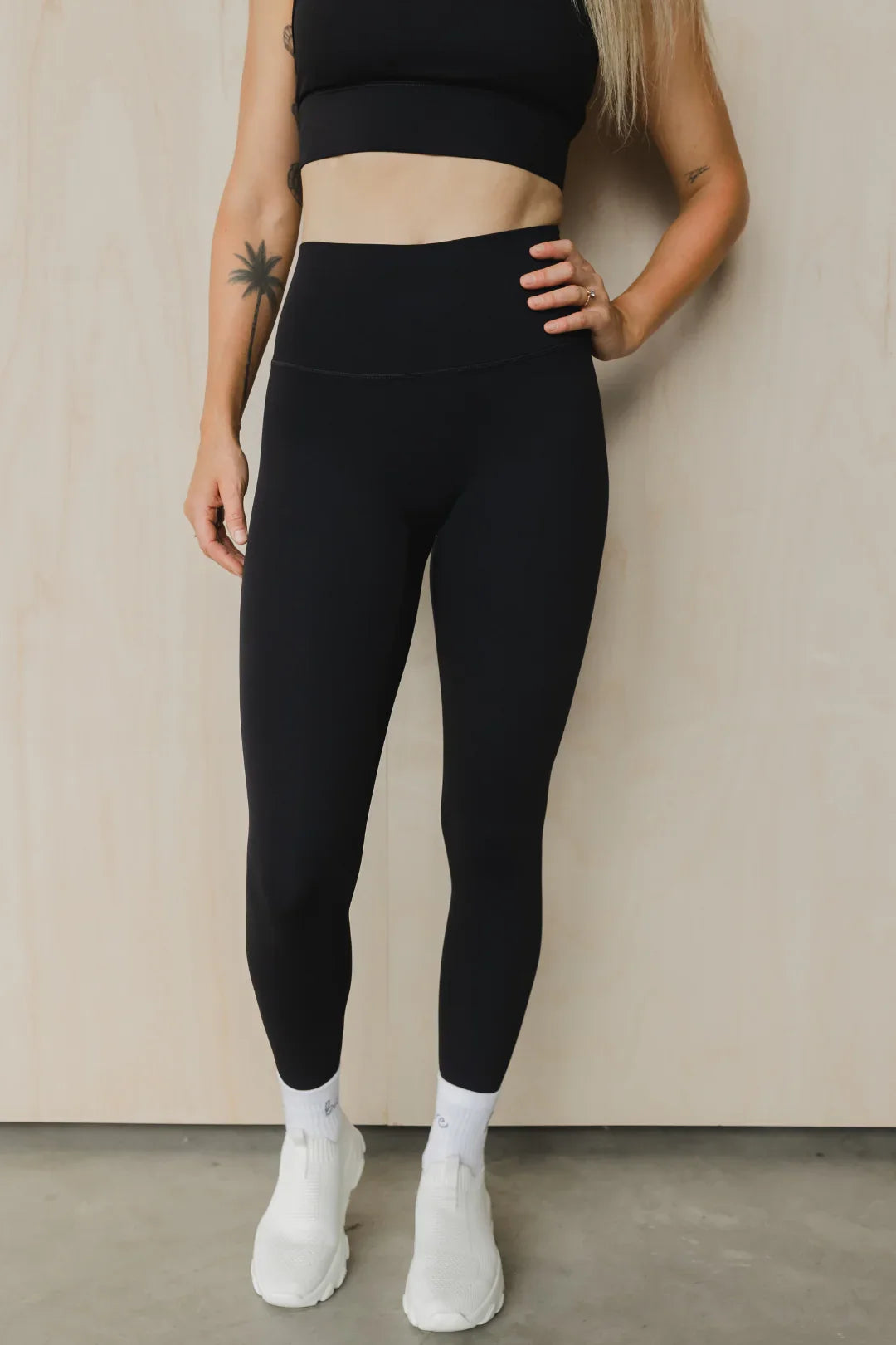 Woman in black high-waisted leggings and crop top, white sneakers, standing indoors