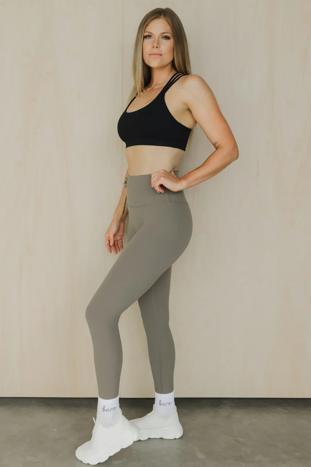 Woman in activewear with black sports bra, taupe leggings, white sneakers on gray floor