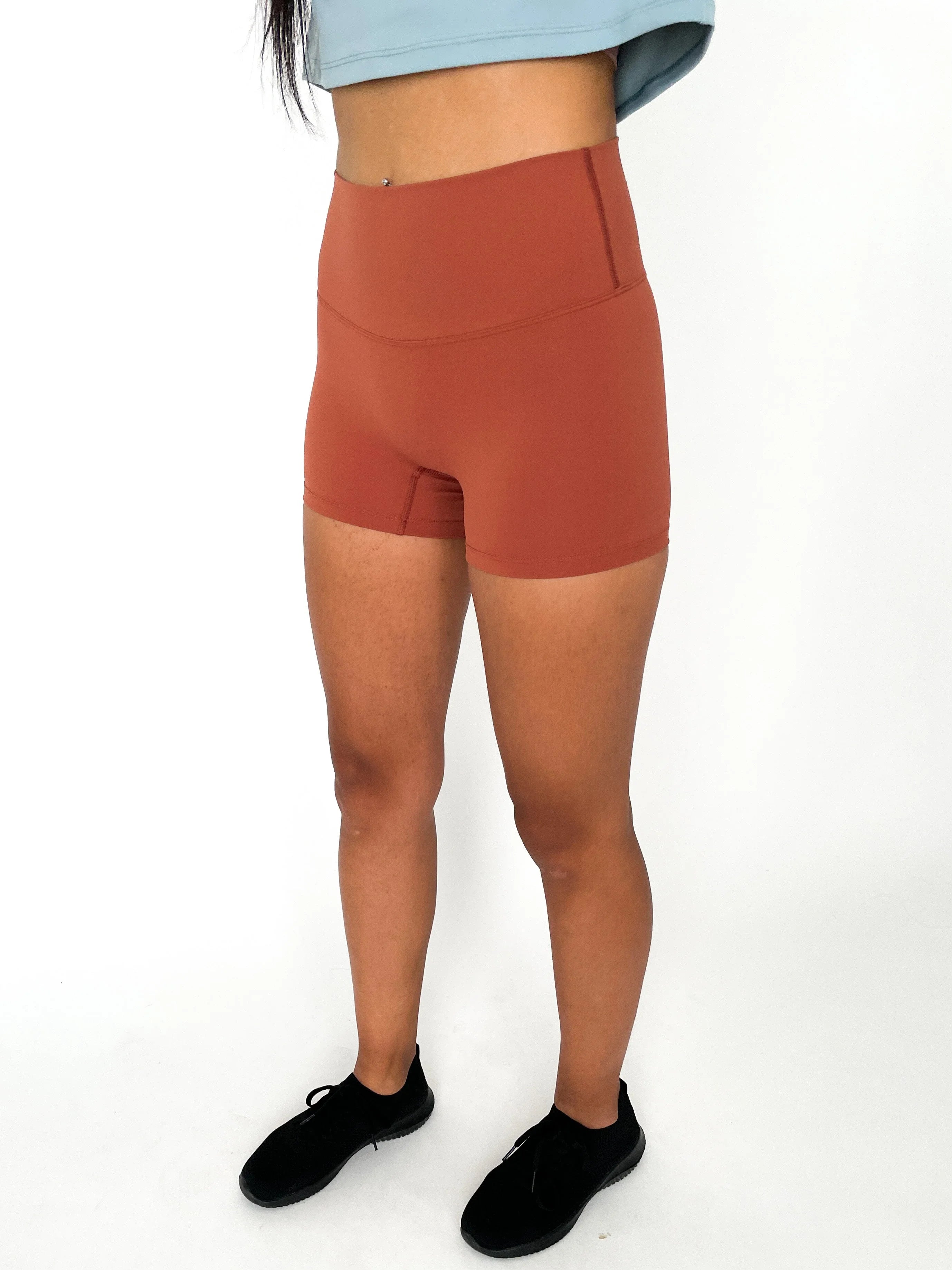 Woman wearing rust high-waisted athletic shorts and black sneakers on white background