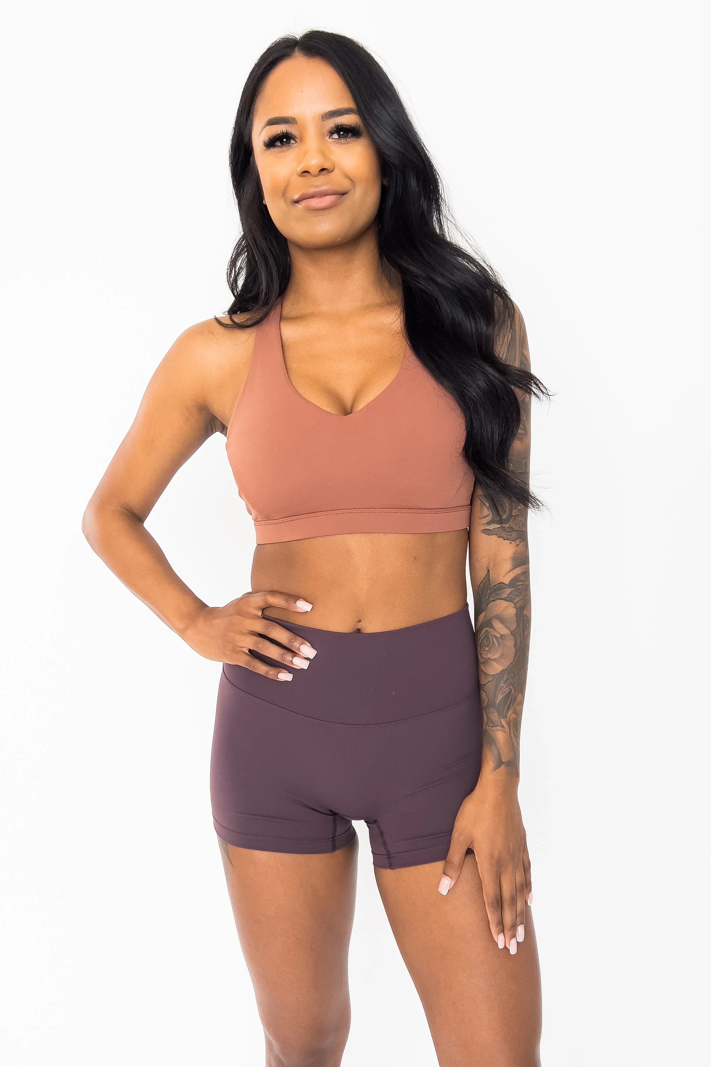 Woman in mauve sports bra and purple workout shorts with floral arm tattoo on white background