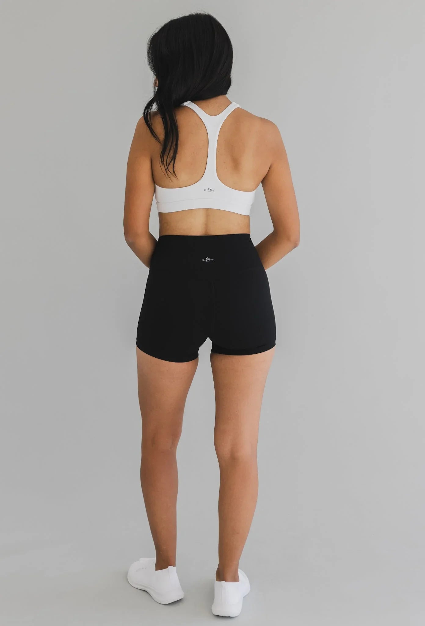 Woman in white sports bra and black high-waisted athletic shorts facing away on gray background