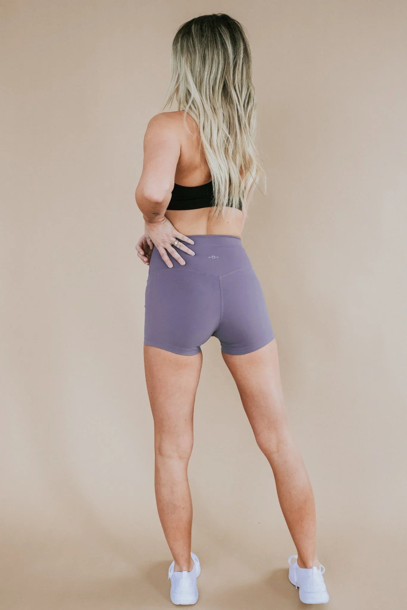 Woman in black sports bra and purple athletic shorts standing, showing back view, studio setting