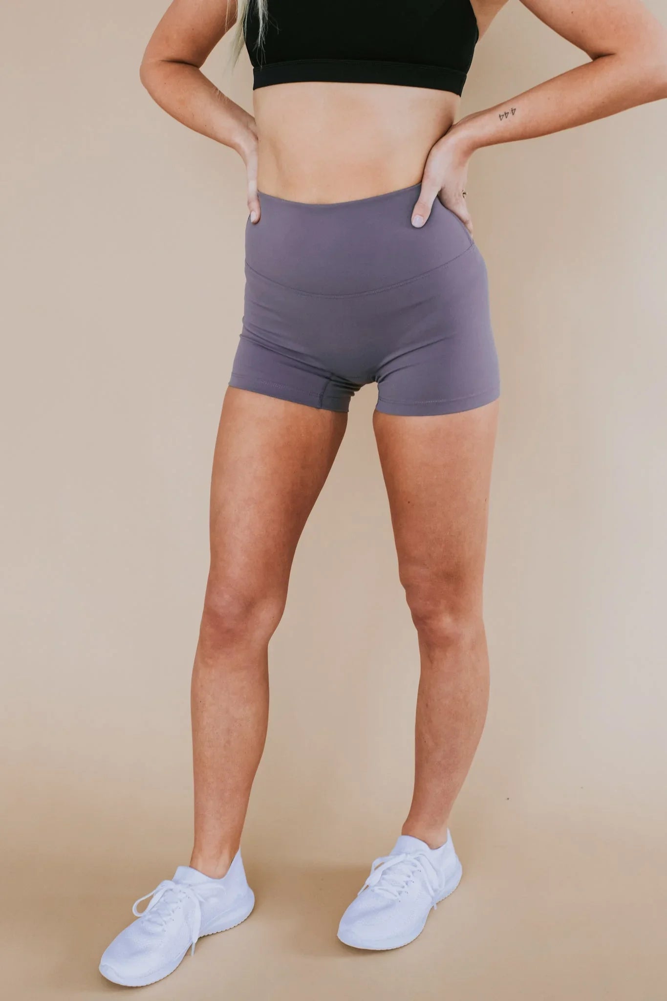 Woman in high-waisted purple athletic shorts, black sports bra, and white sneakers