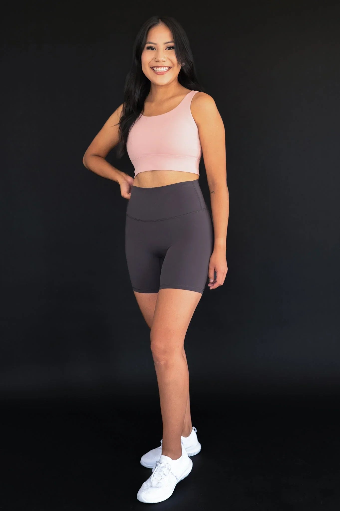 Smiling woman in pink sports bra, gray bike shorts, and white sneakers on black background