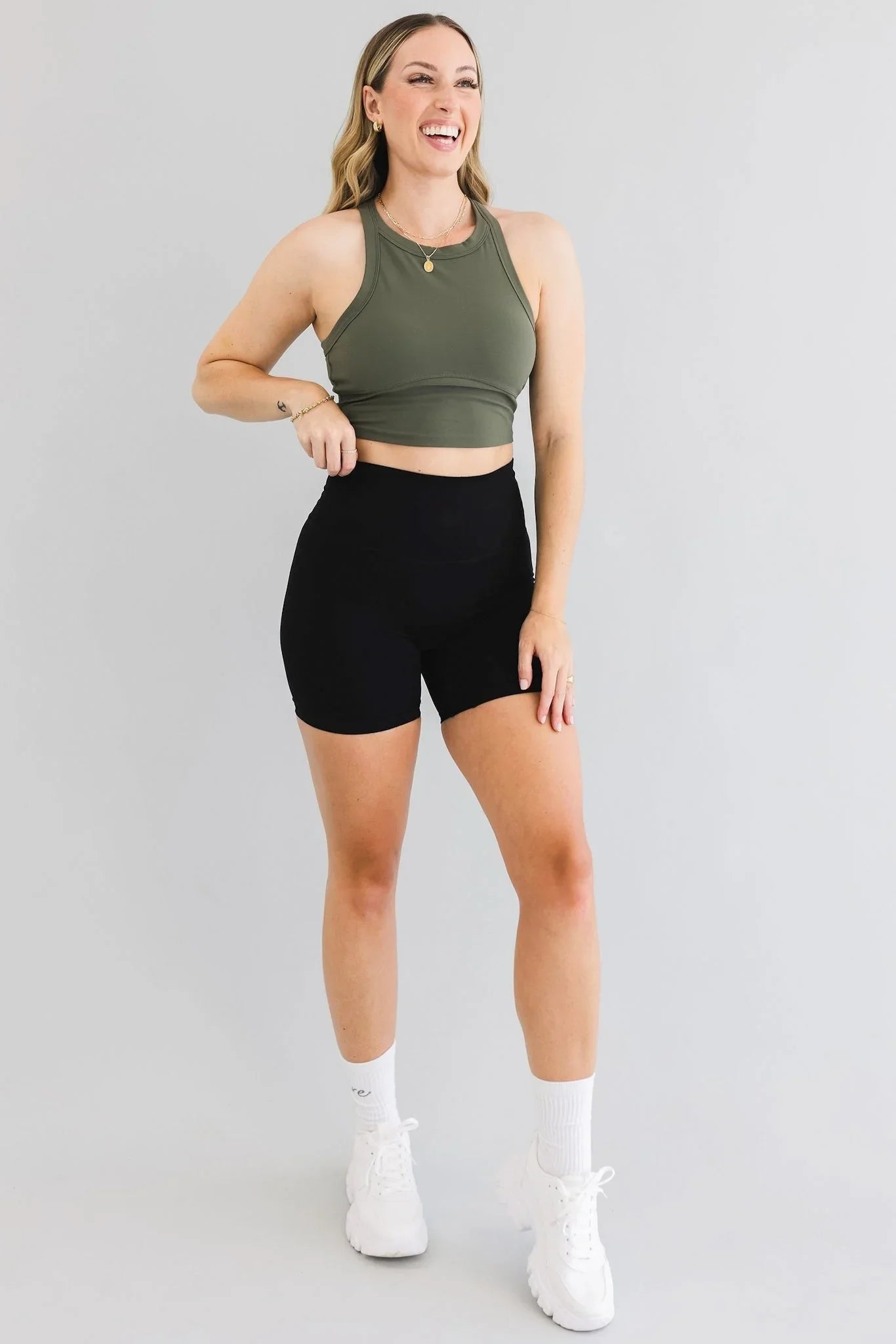 Woman in olive green crop top and black biker shorts, white sneakers, posing against gray background