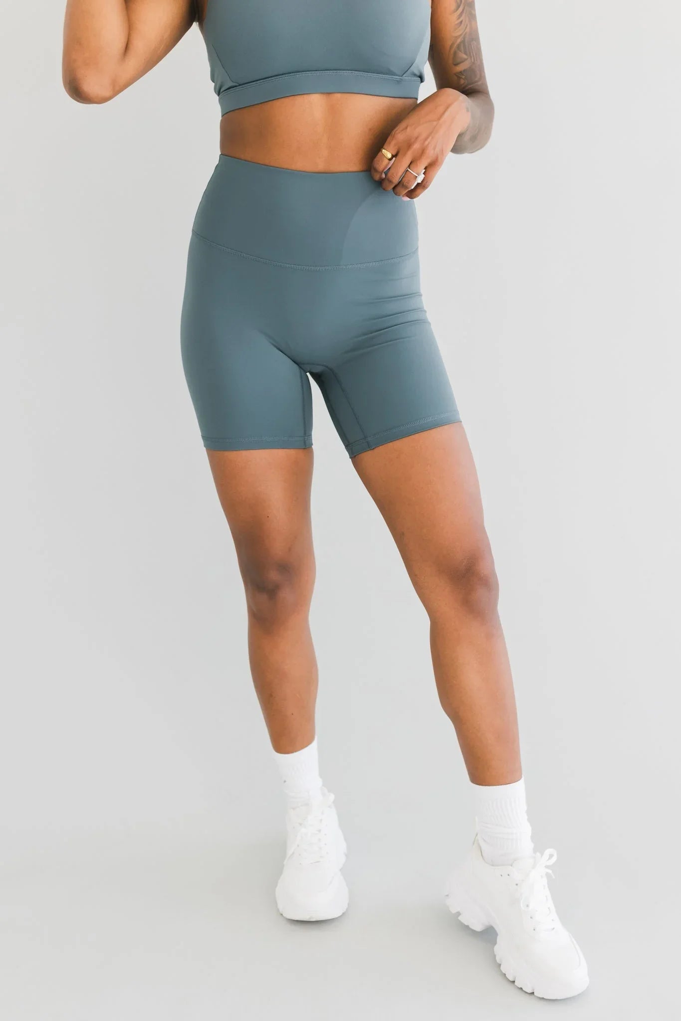 Woman wearing teal high-waisted biker shorts and sports bra, white sneakers, studio background
