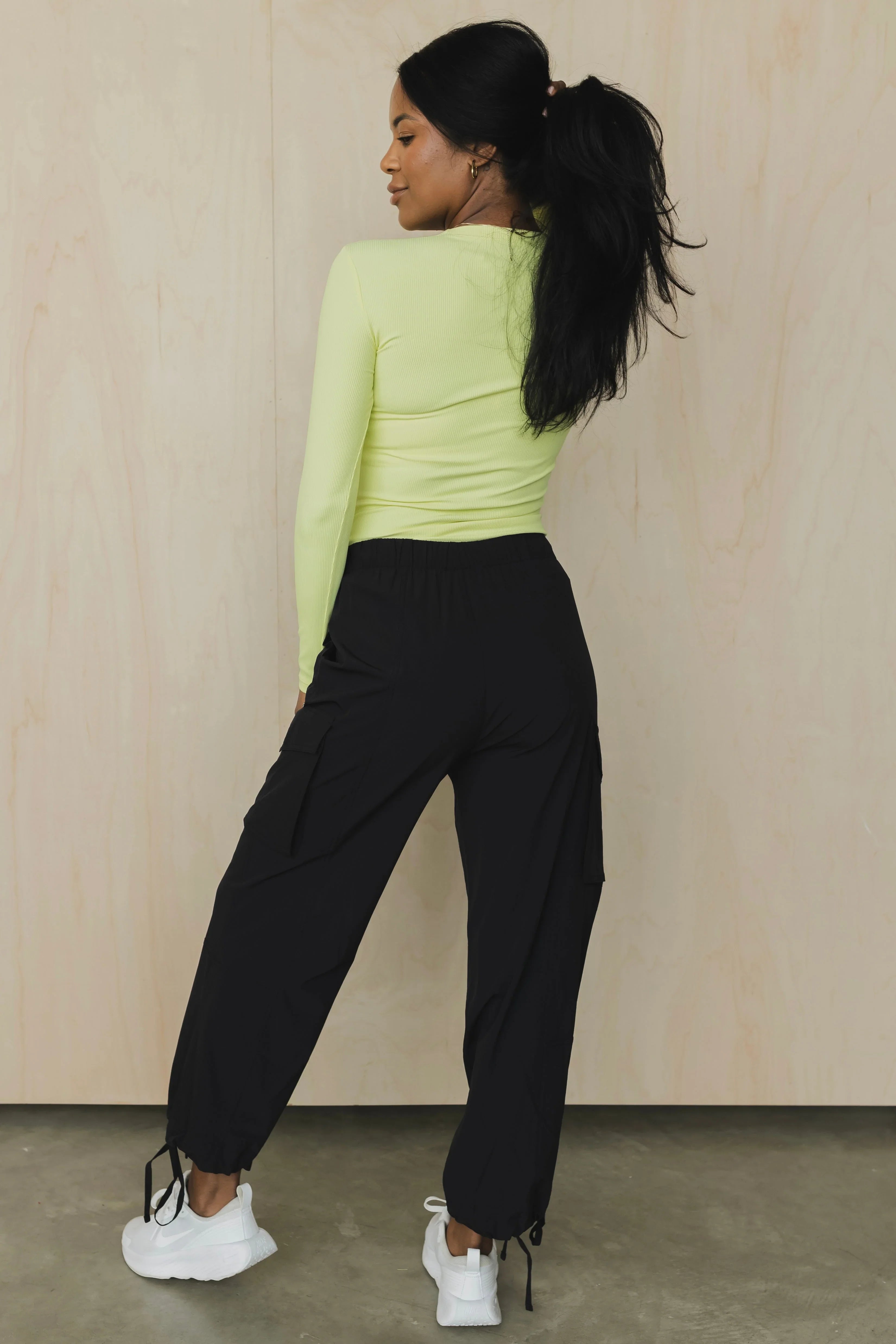 Woman in a neon yellow long sleeve top, black cargo pants, and white sneakers, standing indoors