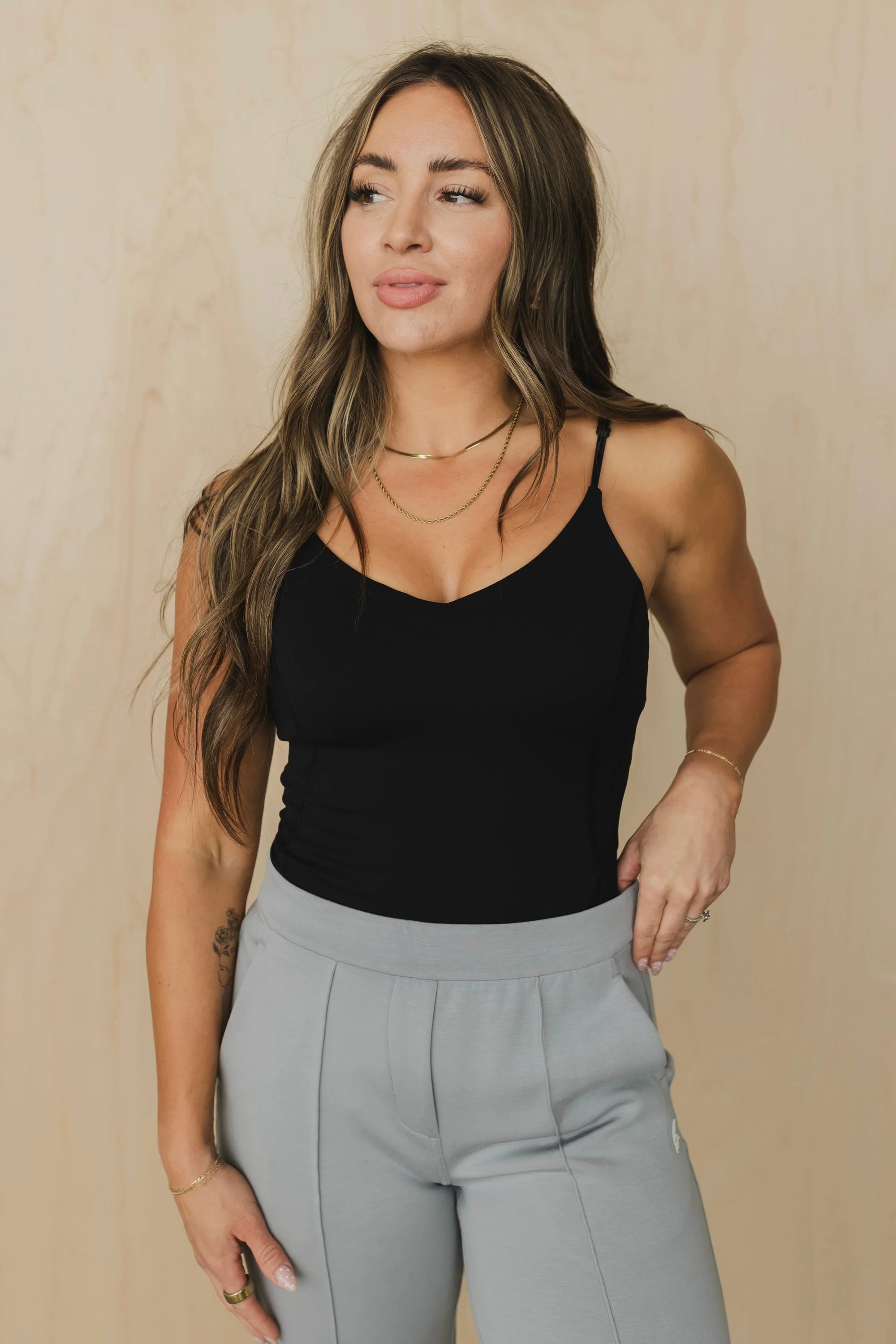 Woman in black camisole top and gray pants with gold jewelry, standing against beige background