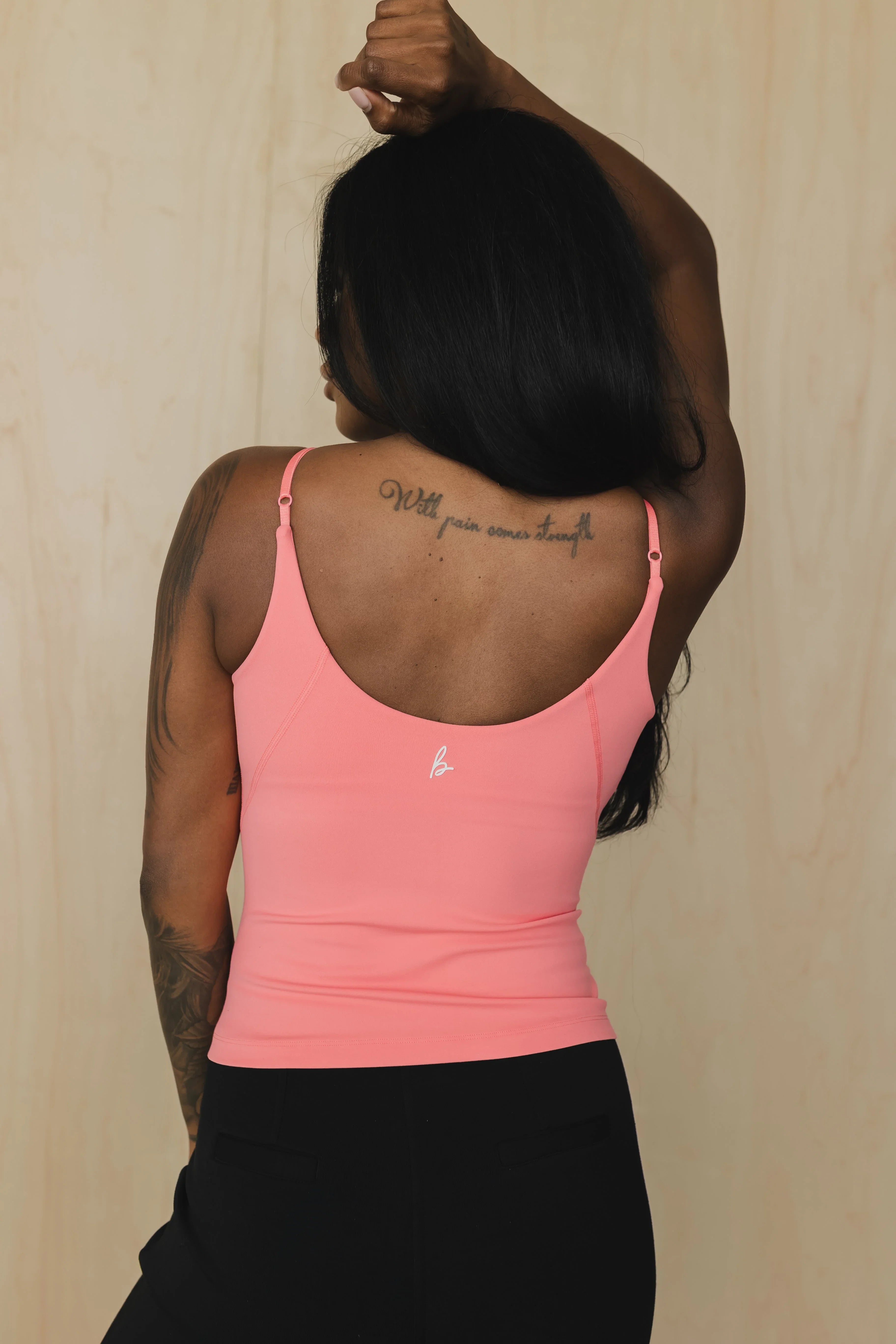 Woman in pink tank top with back tattoo and black pants, posing against a light background.