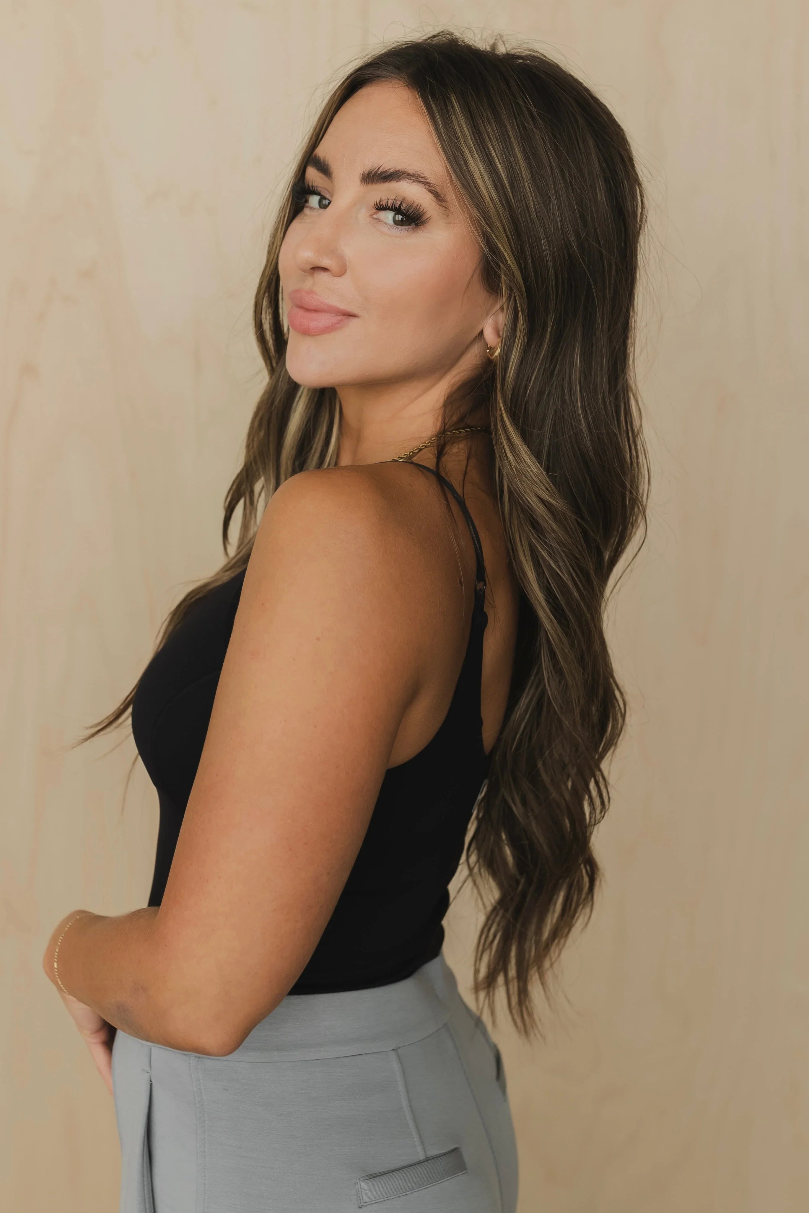 Woman with long wavy brown hair in black top and gray pants, neutral background