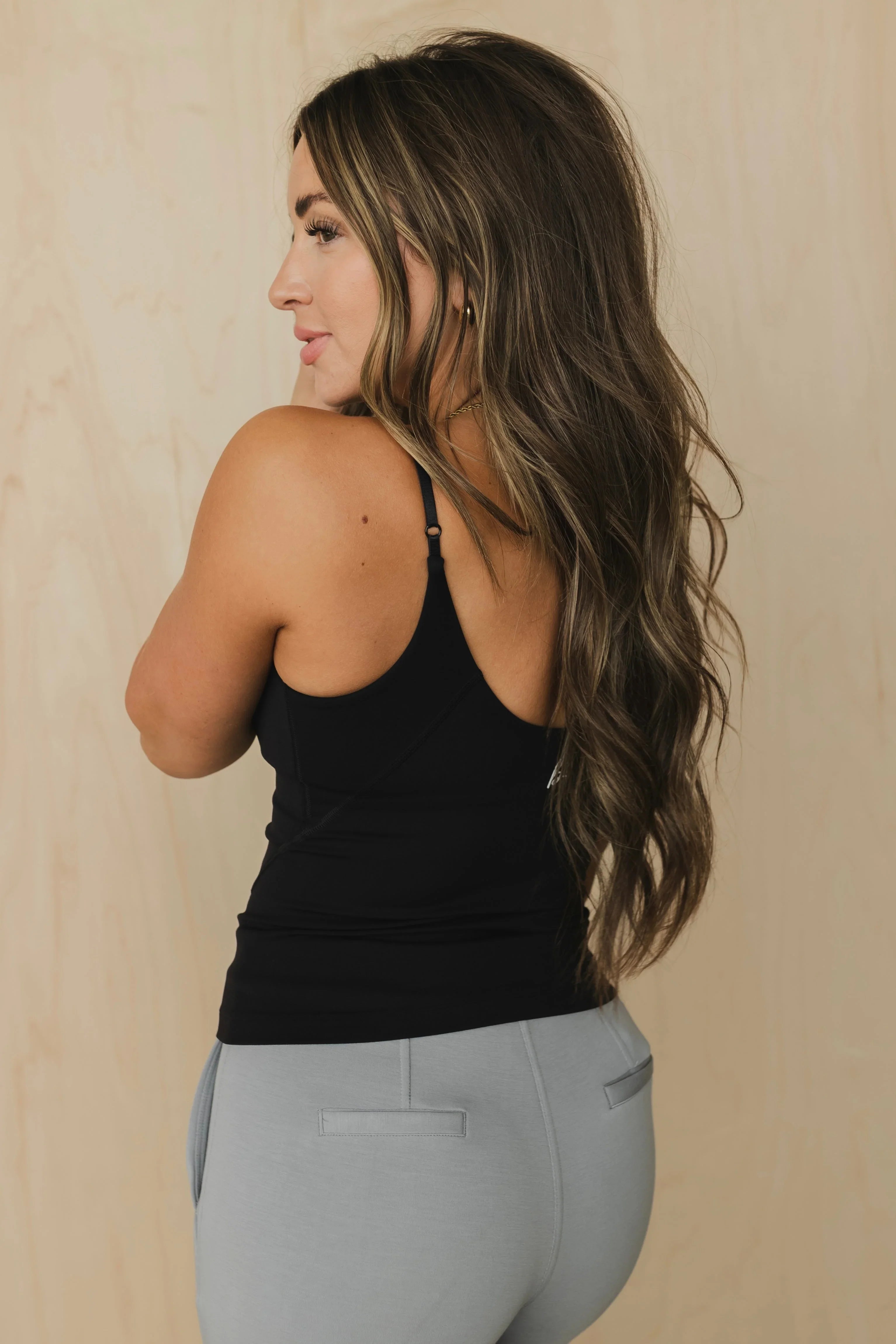 Woman with long wavy brown hair in black tank top and gray pants, neutral background