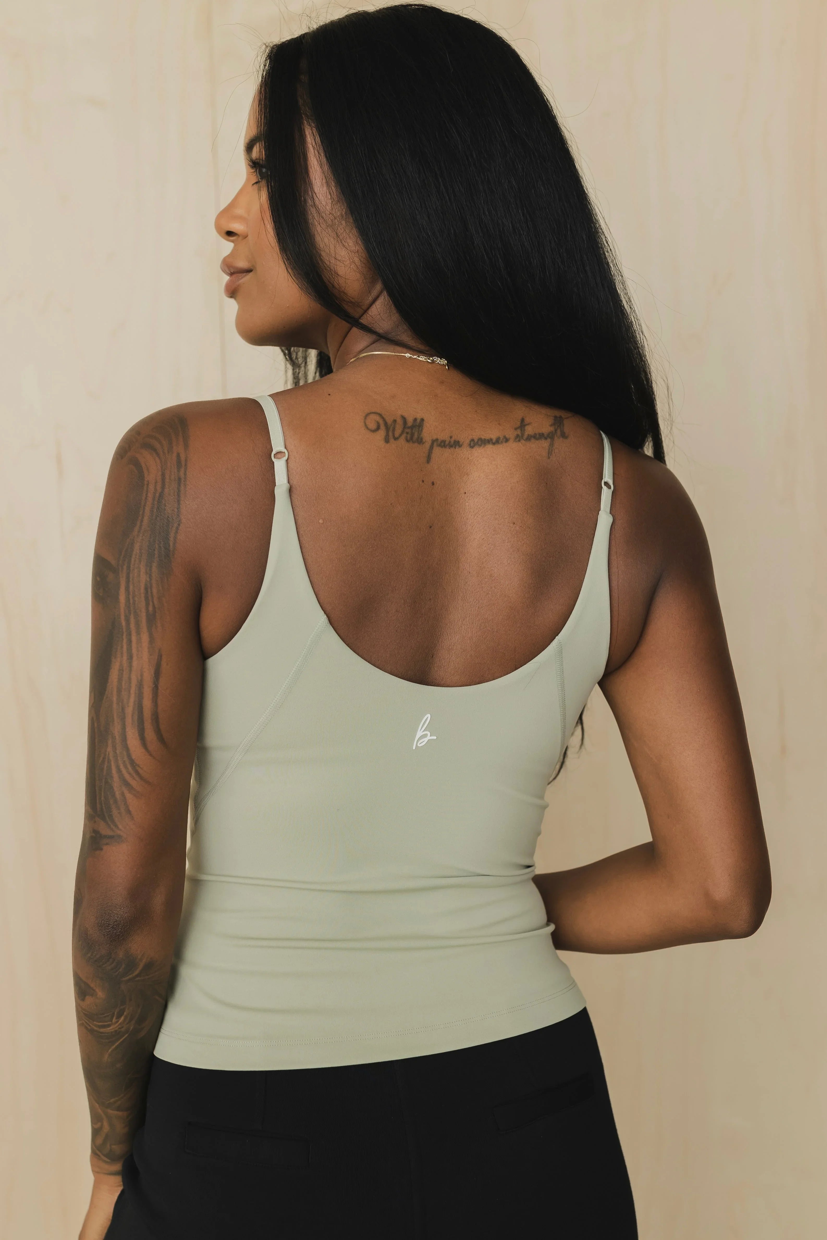 Woman with tattoo and arm sleeve in sage green tank top, black pants, neutral background