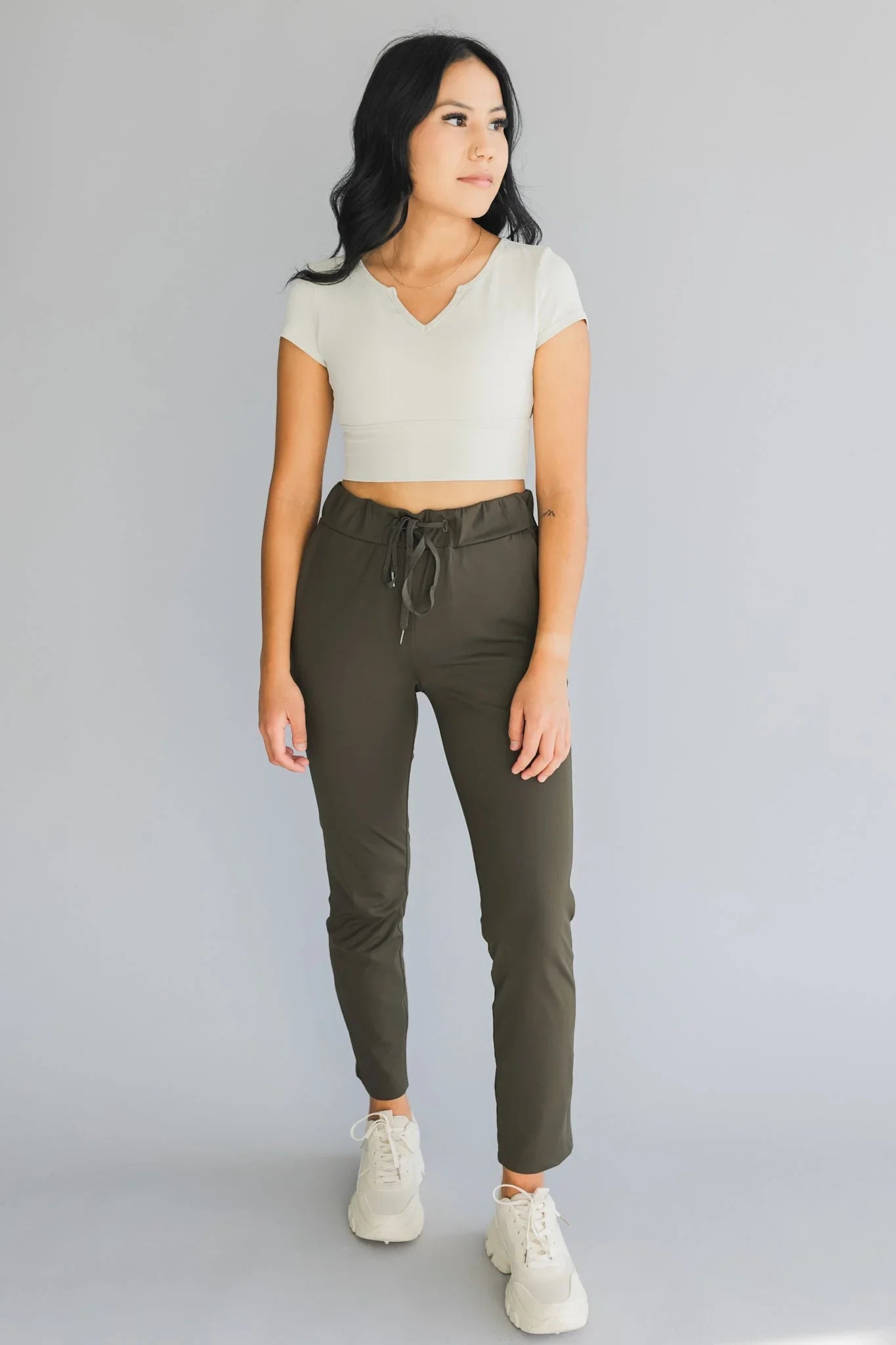 Woman wearing olive jogger pants, white crop top, and sneakers standing against gray background