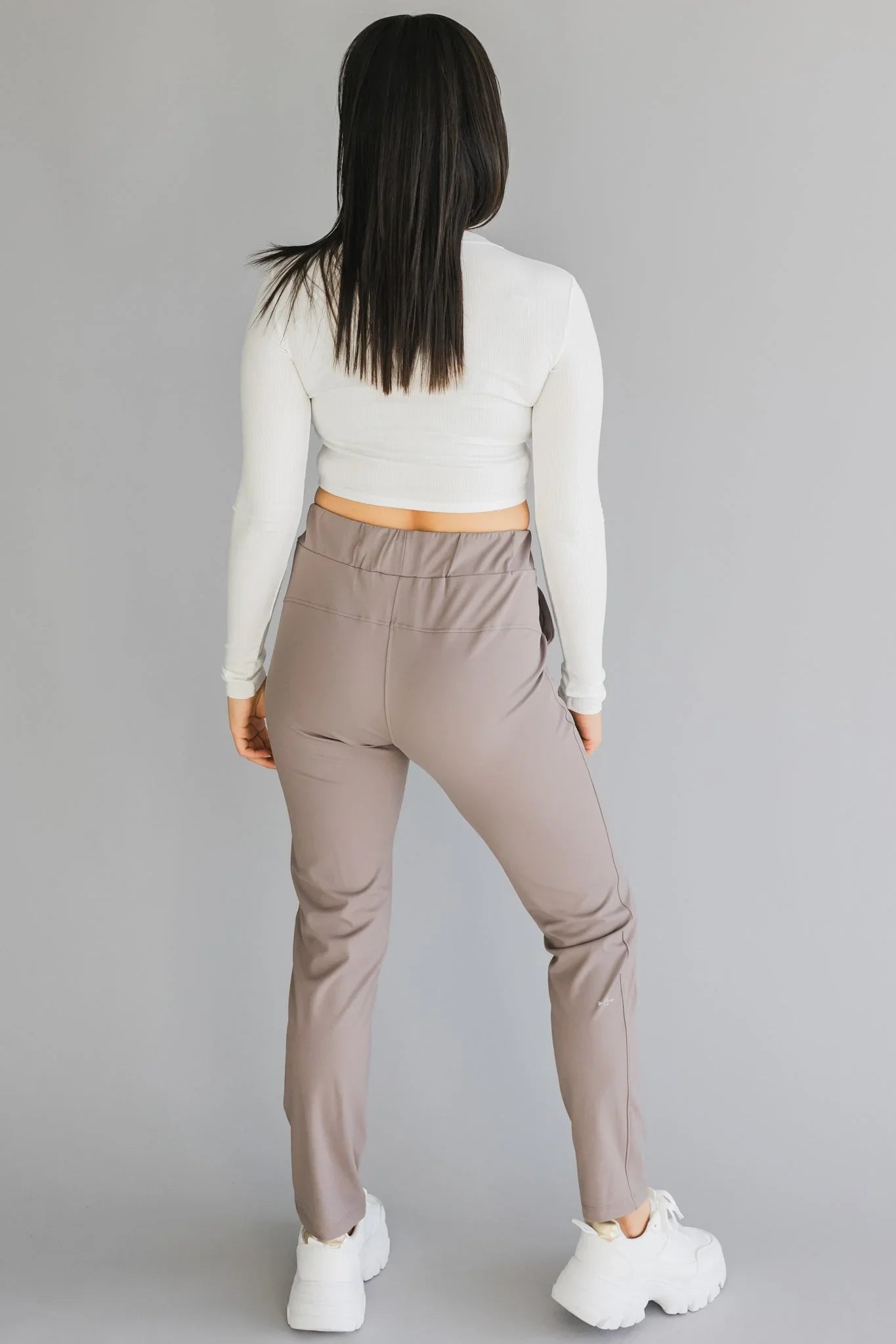 Woman in white long-sleeve crop top and taupe jogger pants, back view, grey background