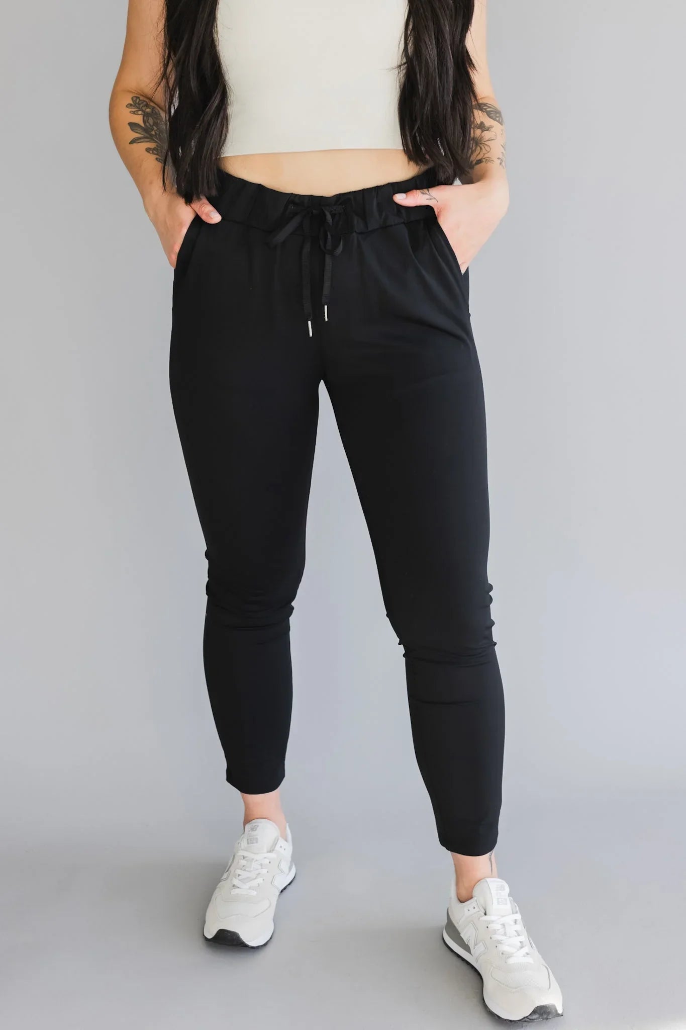 Woman wearing black jogger pants, white crop top, and white sneakers against grey background