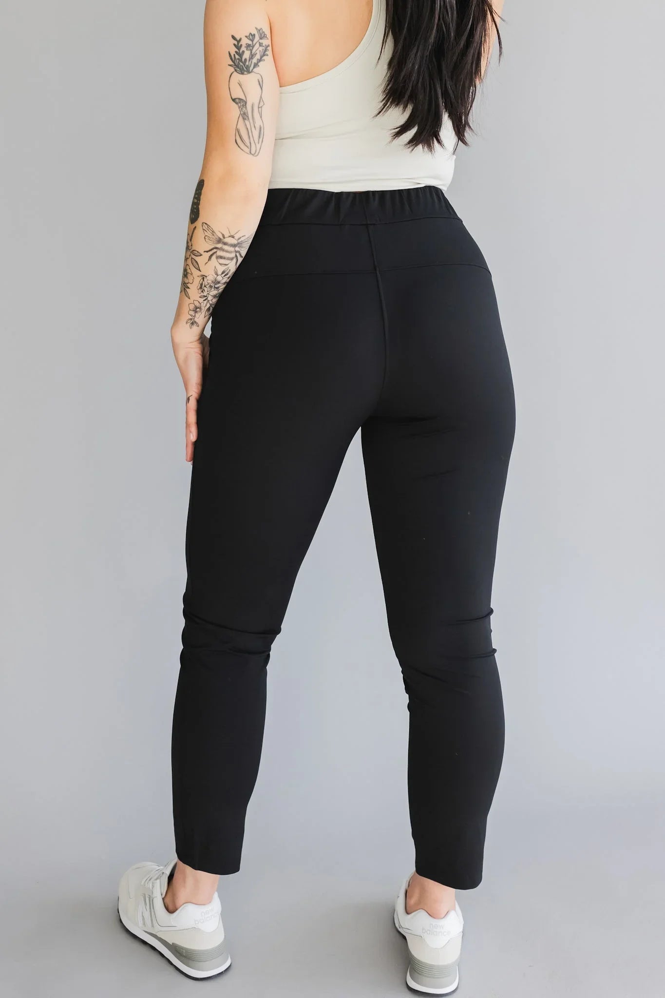 Woman in black leggings, white tank top, white sneakers, tattoos, studio background