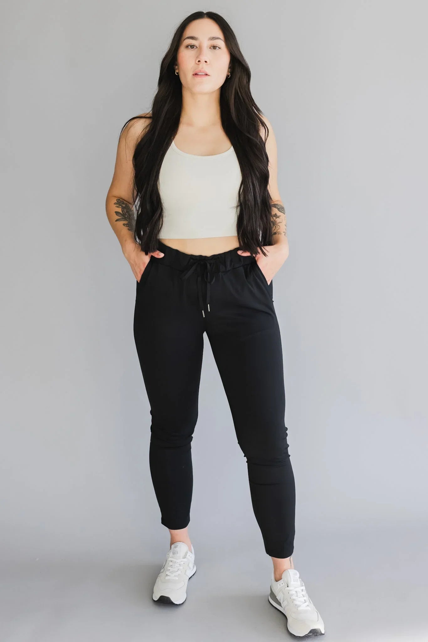 Woman in black joggers, white crop top, and sneakers standing against gray background
