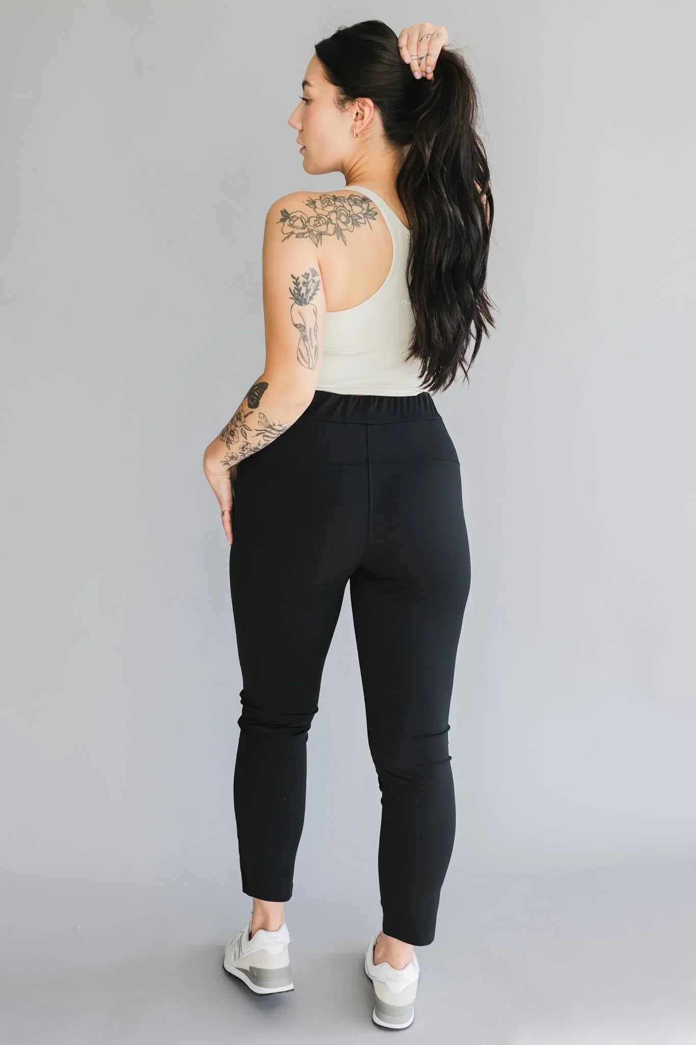 Woman in athletic wear with black leggings, white tank top, and visible arm tattoos stands against gray background