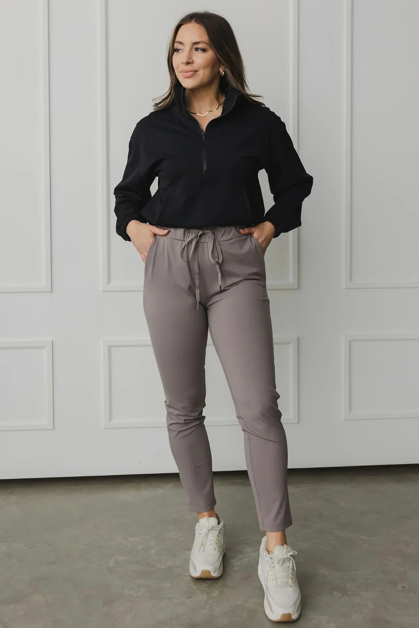 Woman in black zip pullover and taupe jogger pants with white sneakers, posing indoors
