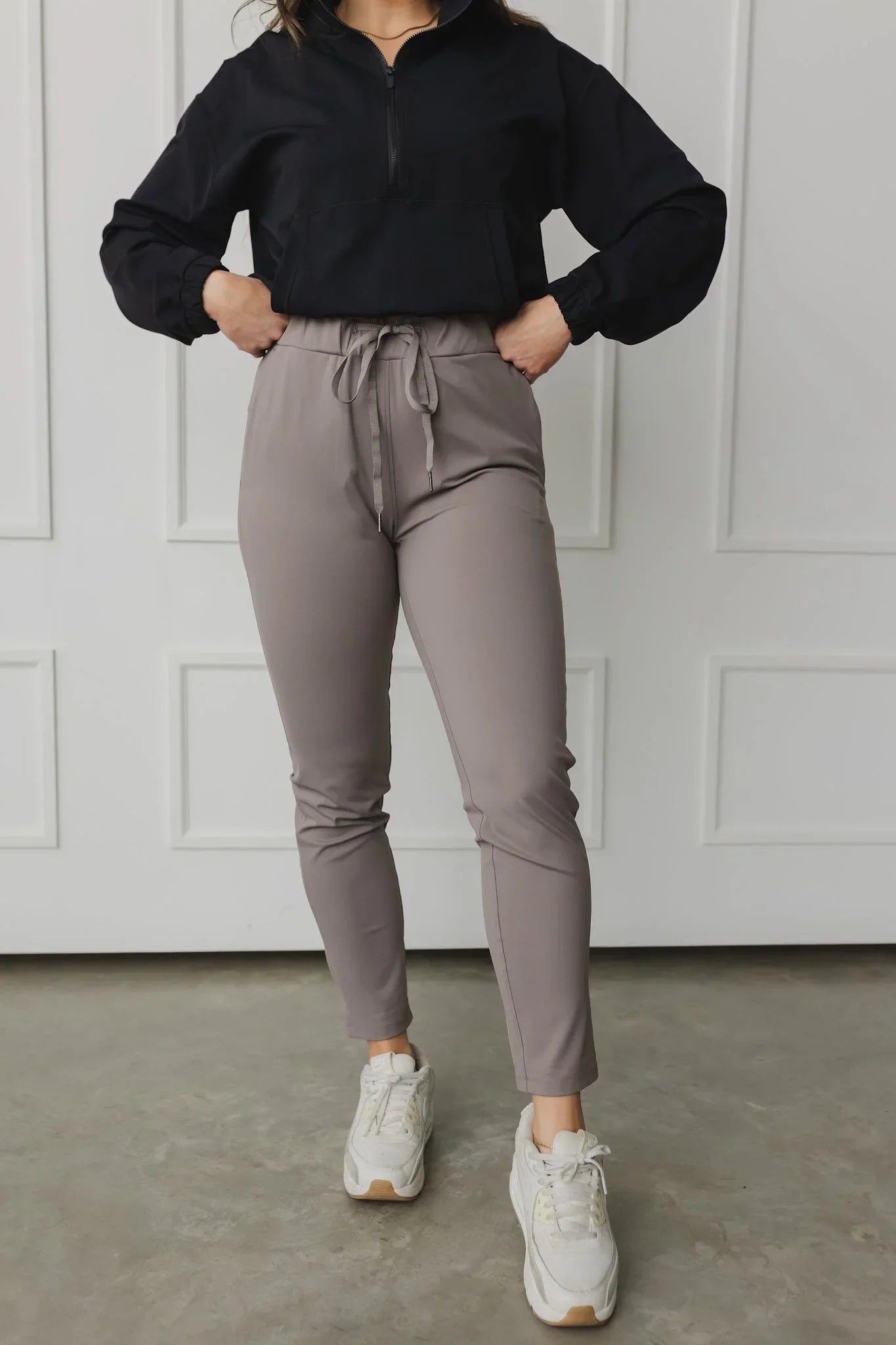 Woman wearing taupe jogger pants with drawstring, black pullover, white sneakers indoors