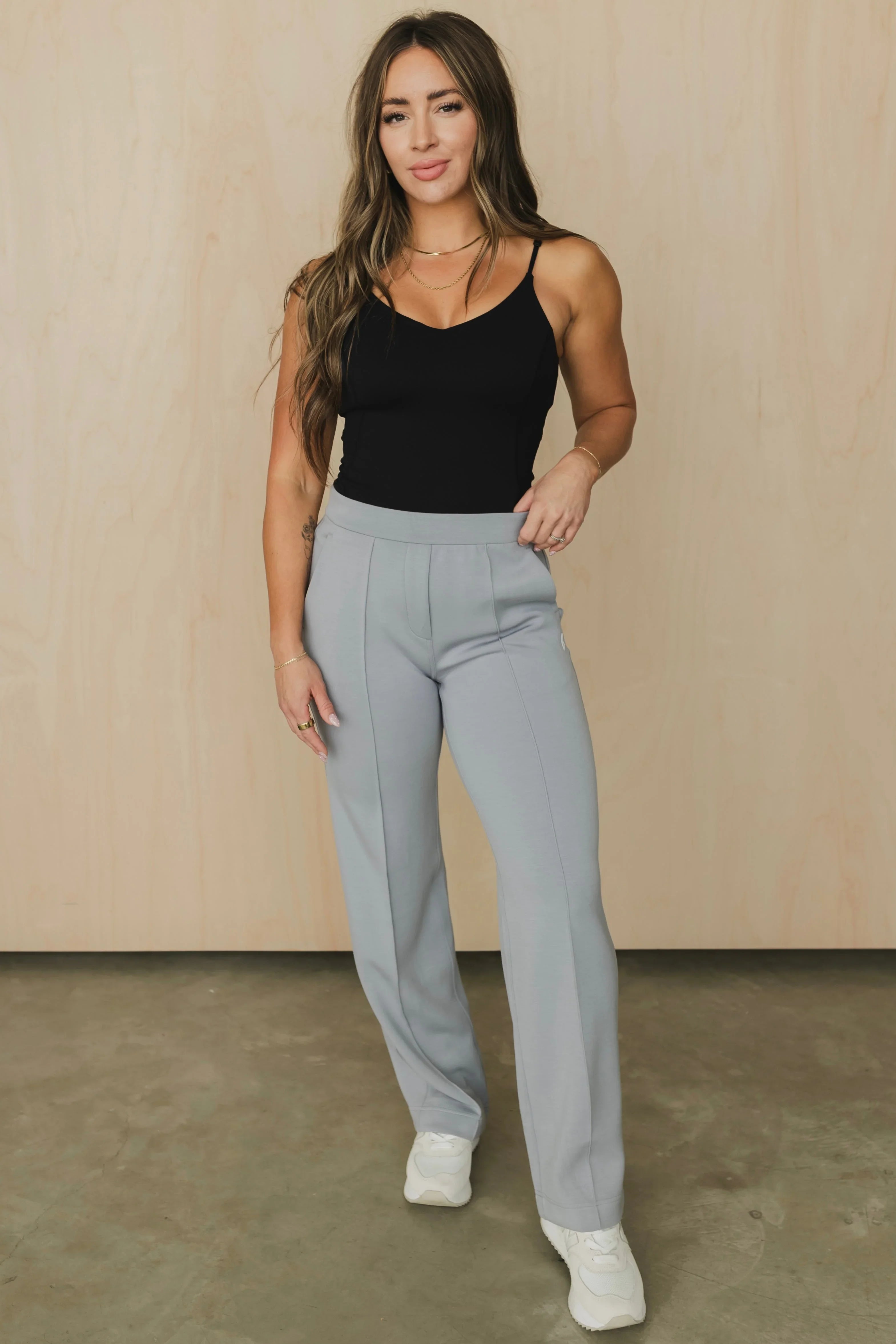 Woman in a black camisole and light gray dress pants with white sneakers standing indoors