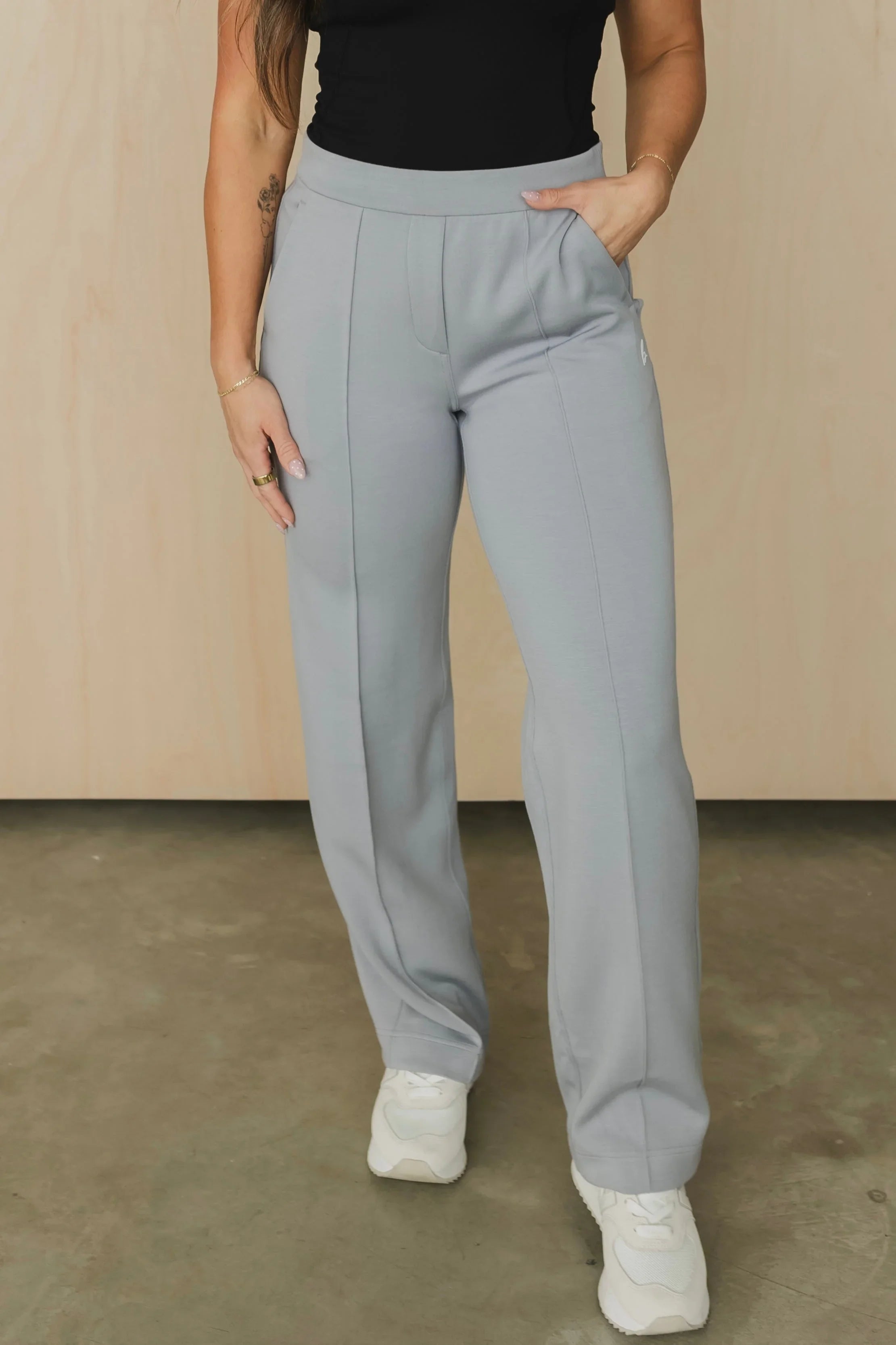 Woman in light gray wide leg pants, black top, and white sneakers standing indoors