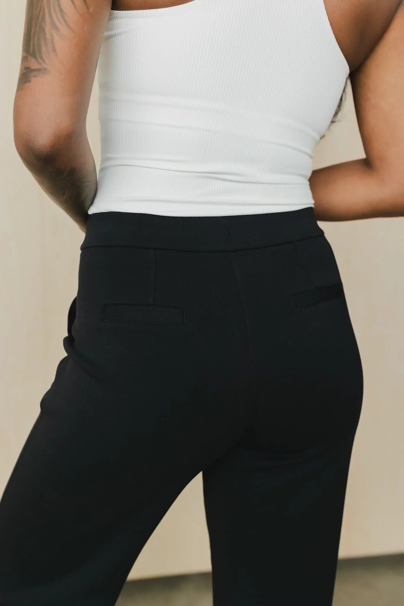 Woman in white ribbed tank top and black pants with tattooed arm, back view