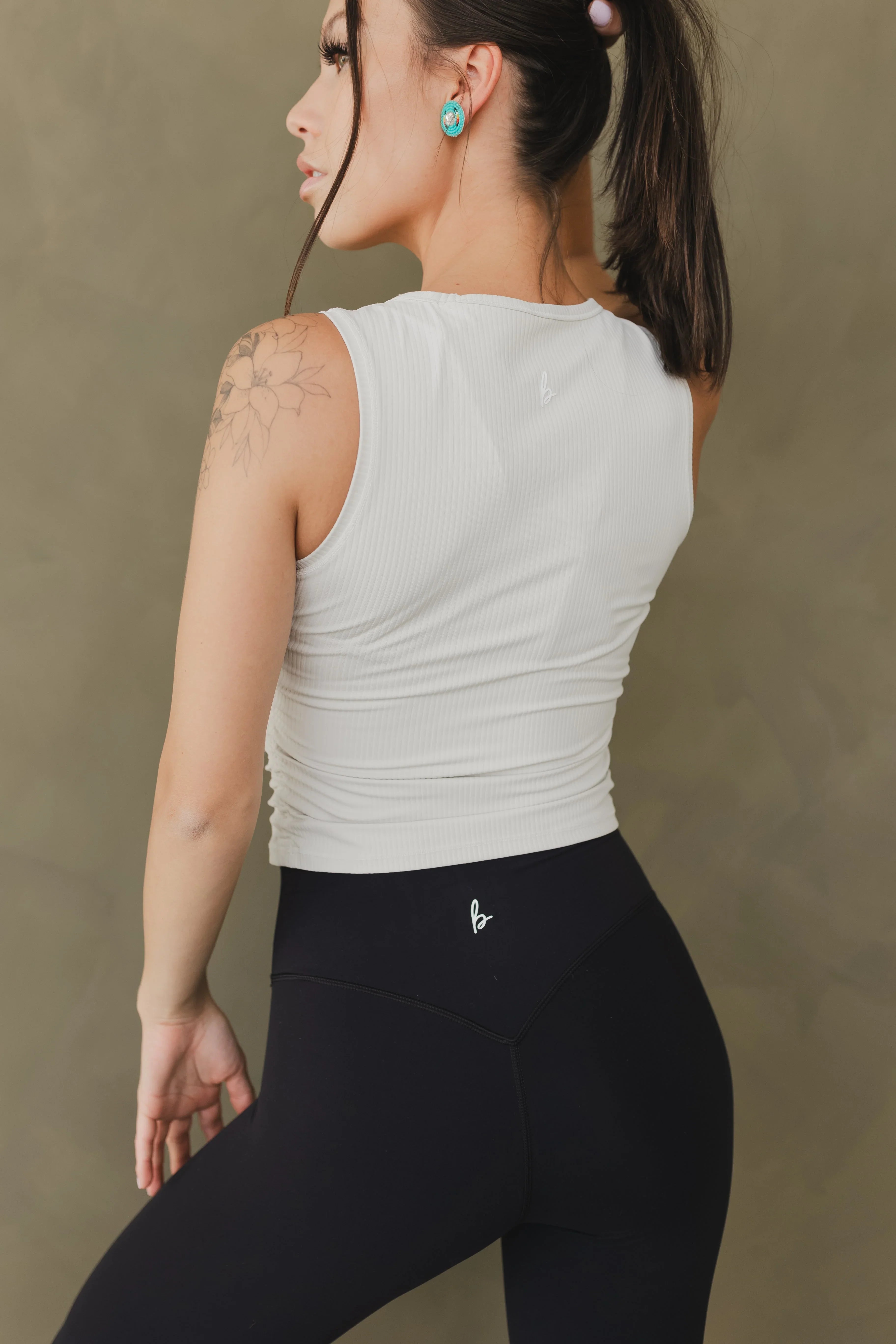 Woman in white ribbed sleeveless top and black leggings with logo, tattoo on shoulder