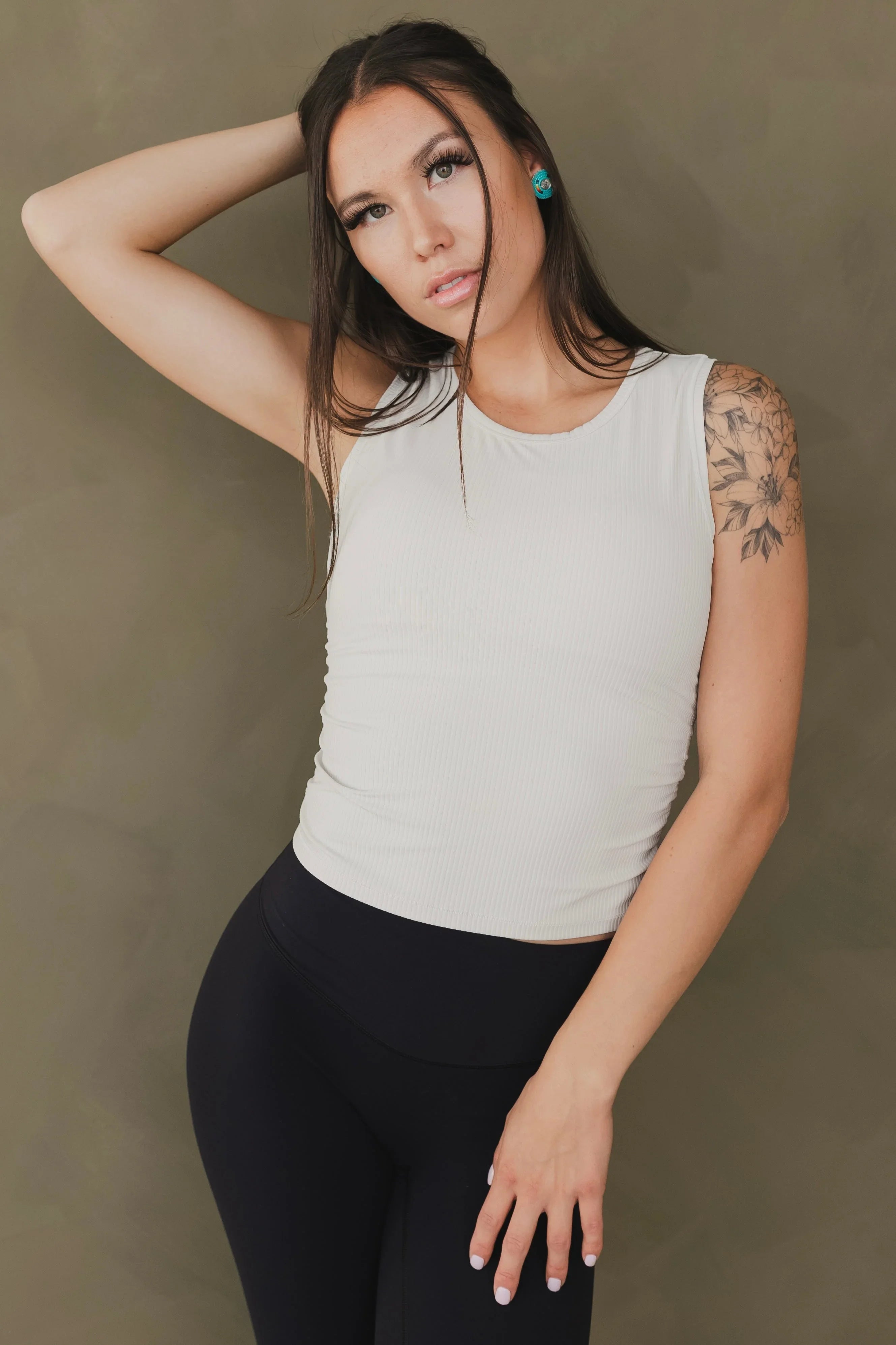 Woman in white sleeveless ribbed top, black leggings, floral arm tattoo, olive background