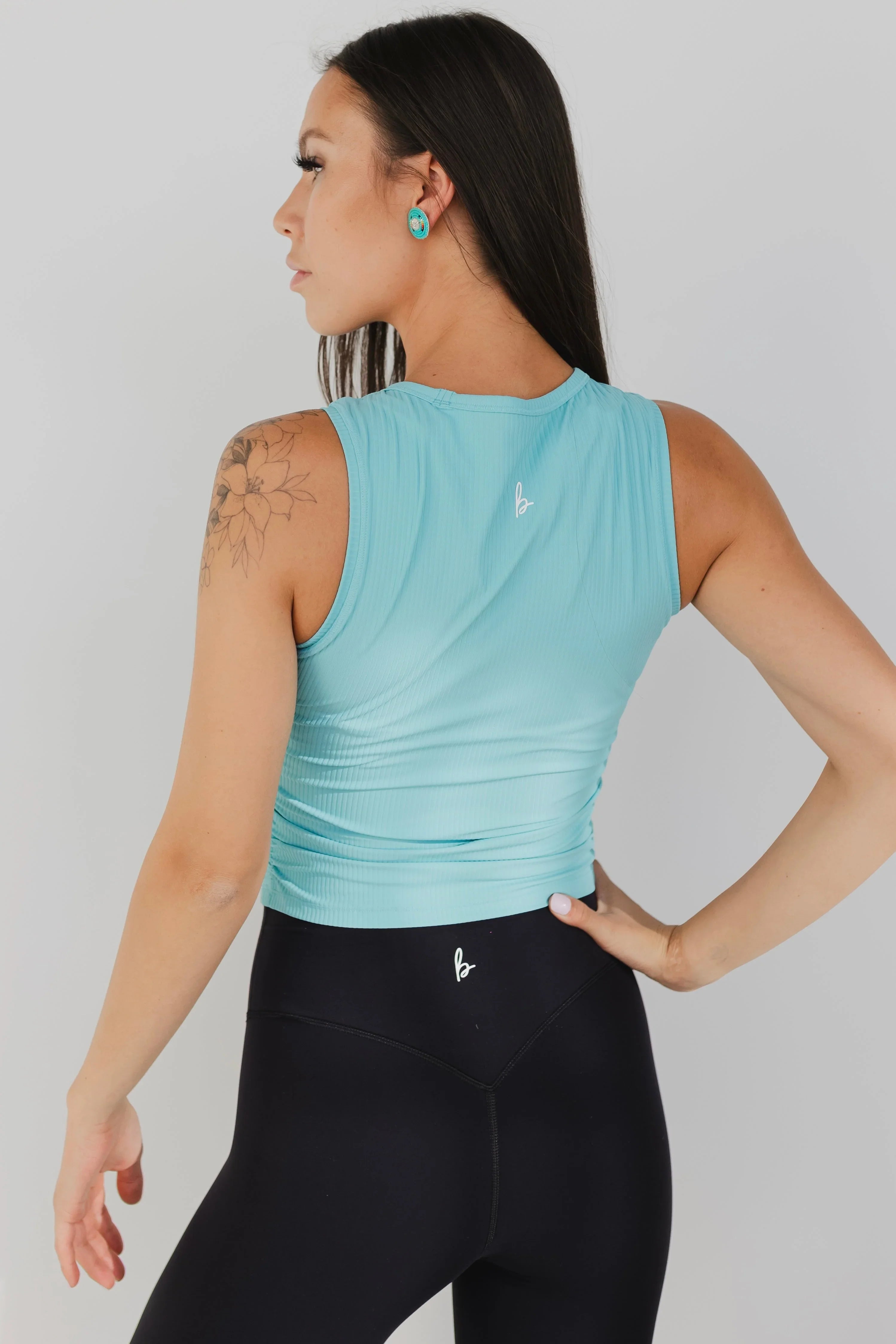 Woman in aqua blue sleeveless top and black leggings, floral arm tattoo, studio background
