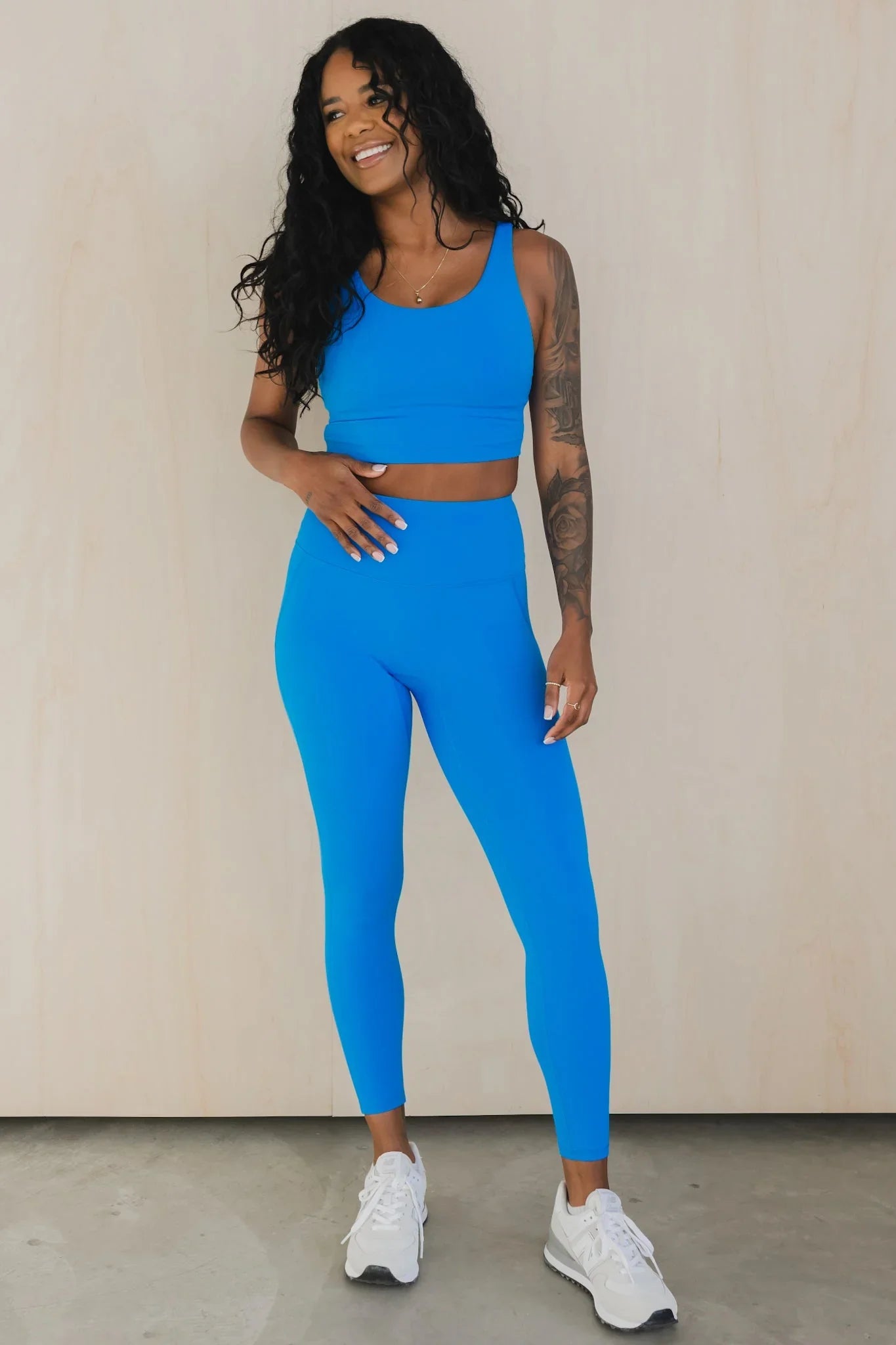 Smiling woman in bright blue sports bra and leggings, white sneakers, standing indoors