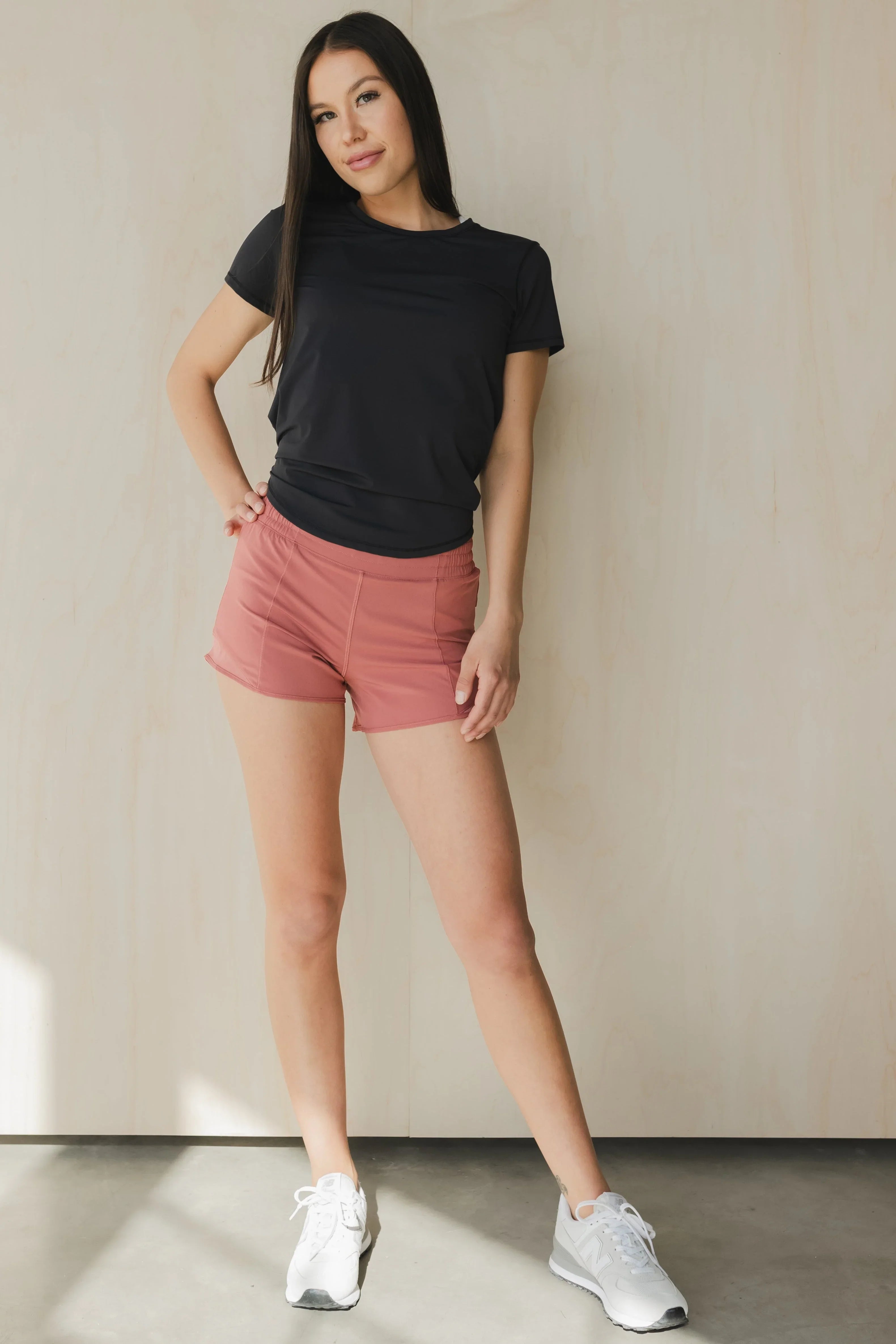 Woman wearing black athletic t-shirt, pink shorts, white sneakers, standing indoors