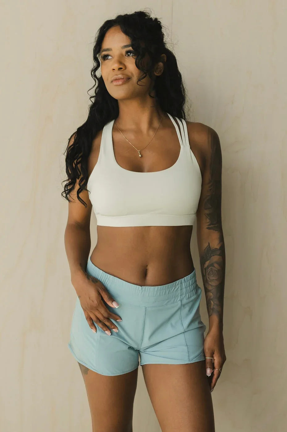 Woman in light sports bra and blue athletic shorts posing against beige wall