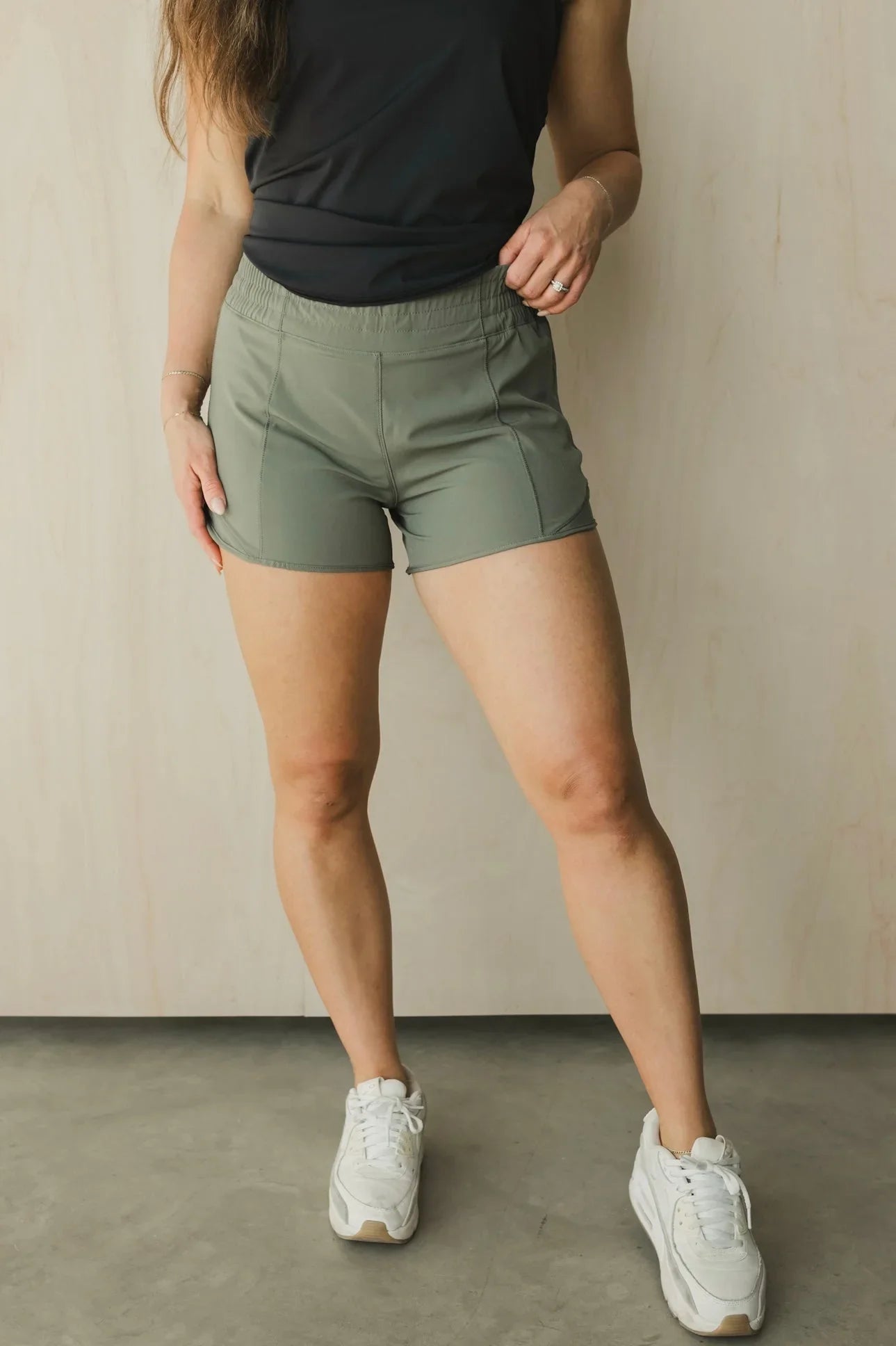 Woman wearing olive green athletic shorts, black tank top, and white sneakers indoors.