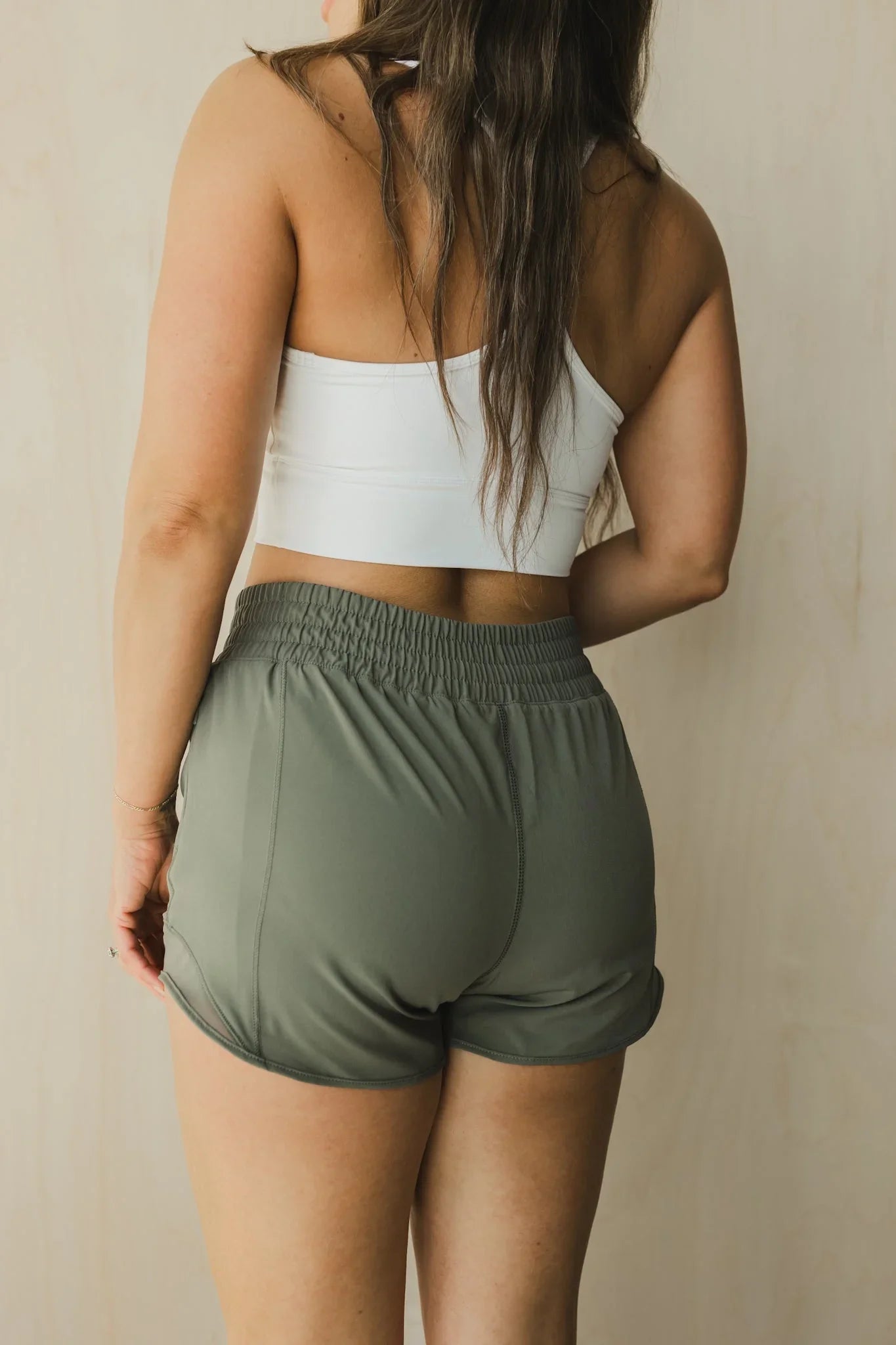Woman in white sports bra and olive green athletic shorts, shown from behind, indoor setting