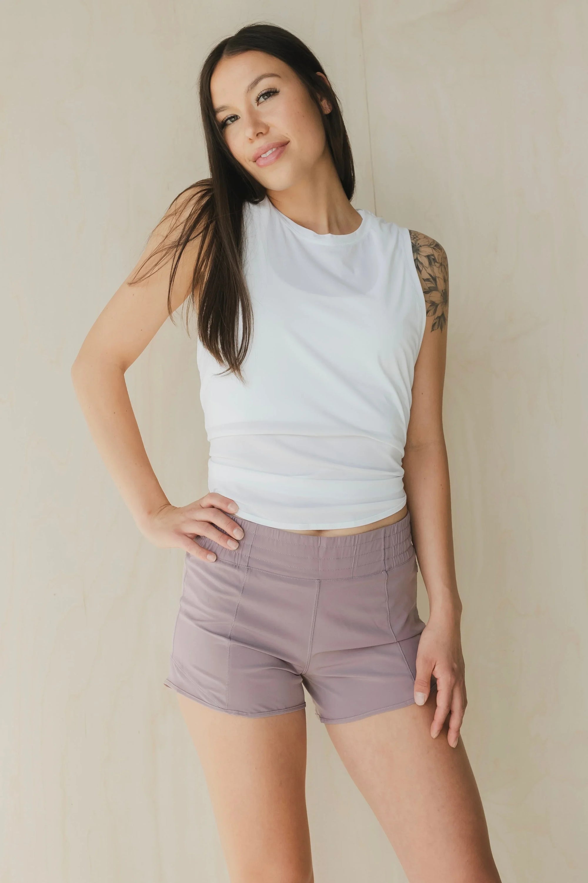 Woman in white sleeveless top and mauve athletic shorts posing against beige wall