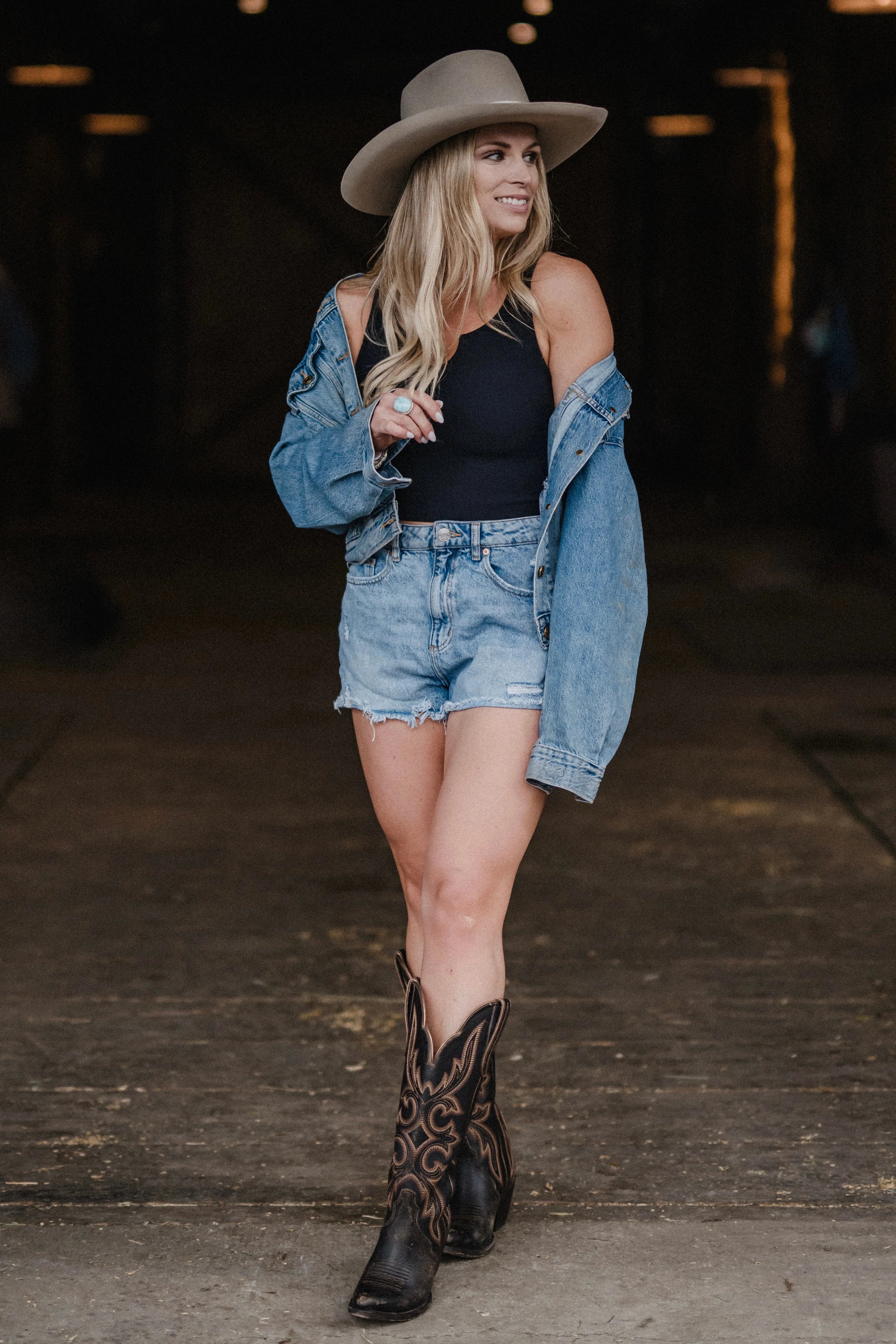 Woman in cowboy hat, denim jacket, black tank, and boots in rustic barn setting