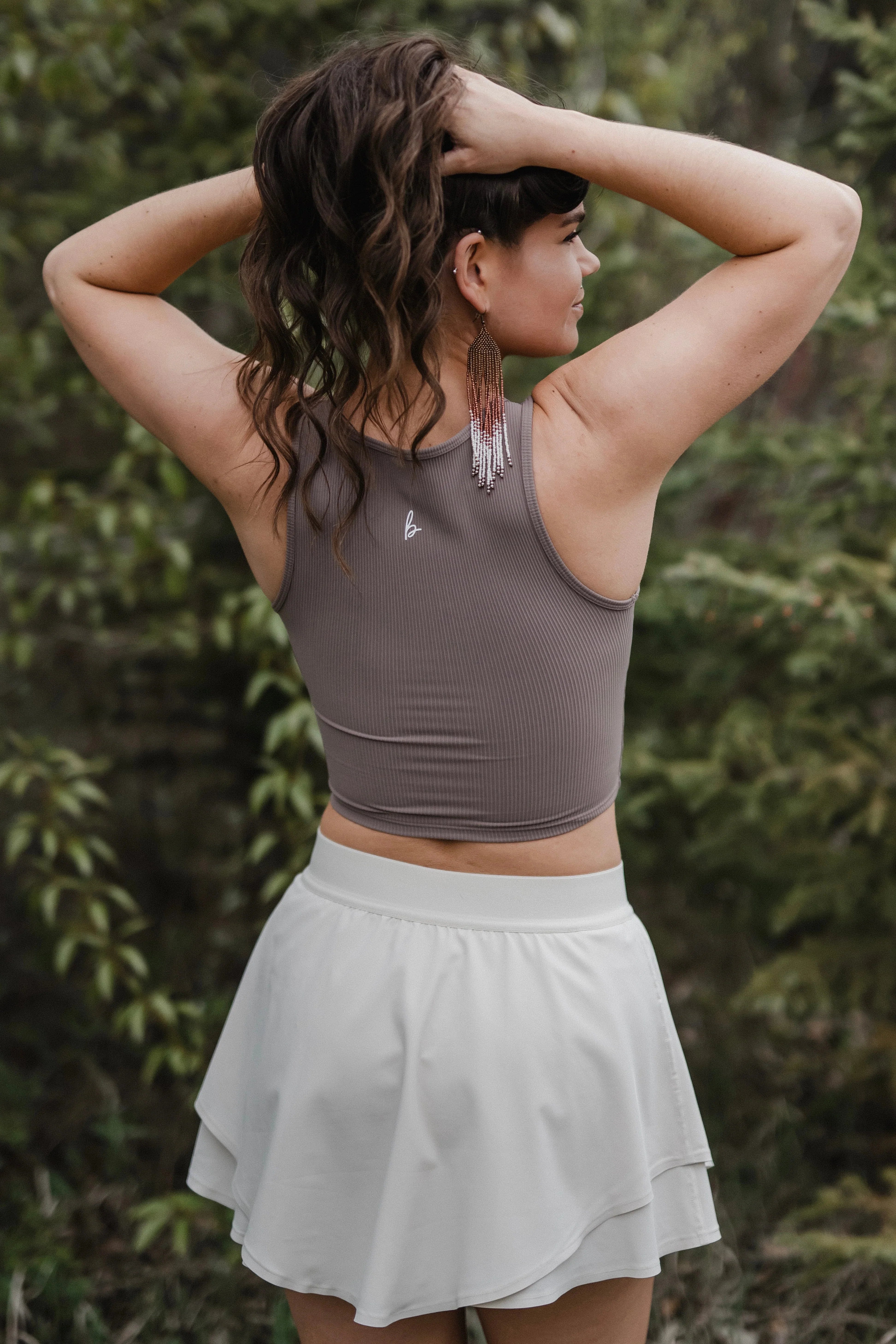 Woman outdoors in taupe crop top, white tennis skirt, and long beaded earrings with greenery background