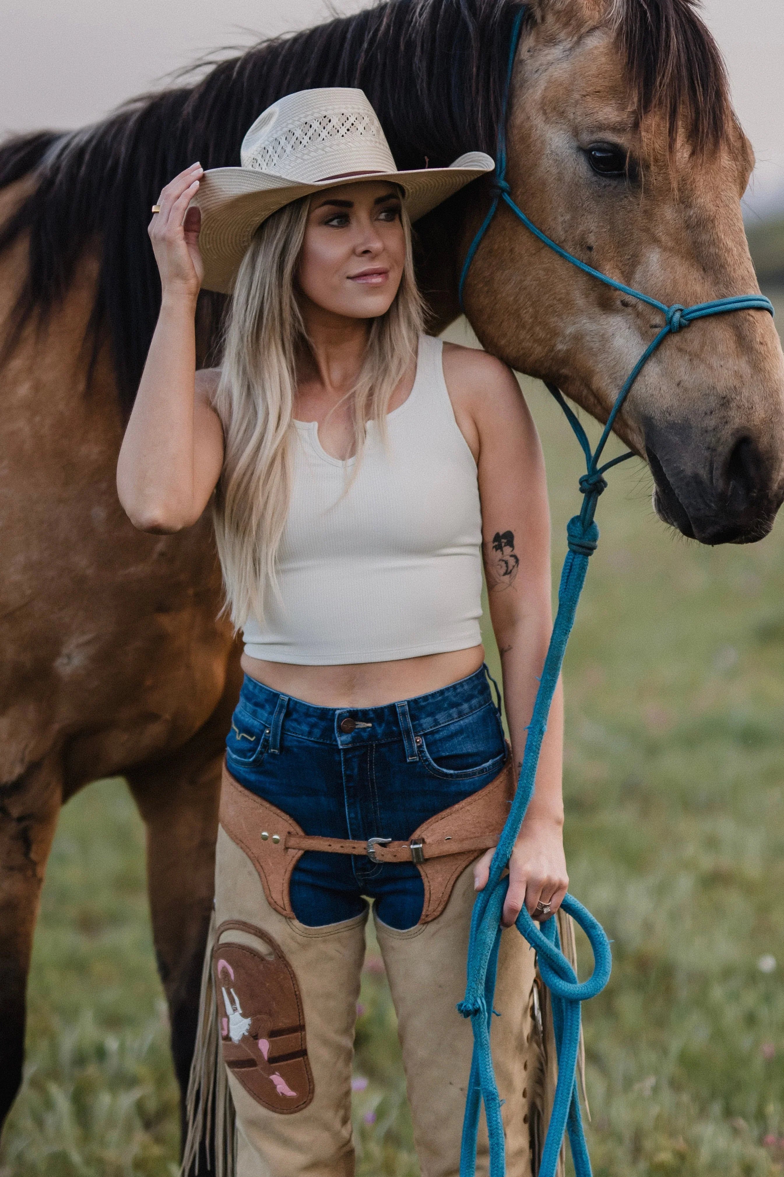Cowgirl in cowboy hat and chaps standing beside brown horse outdoors