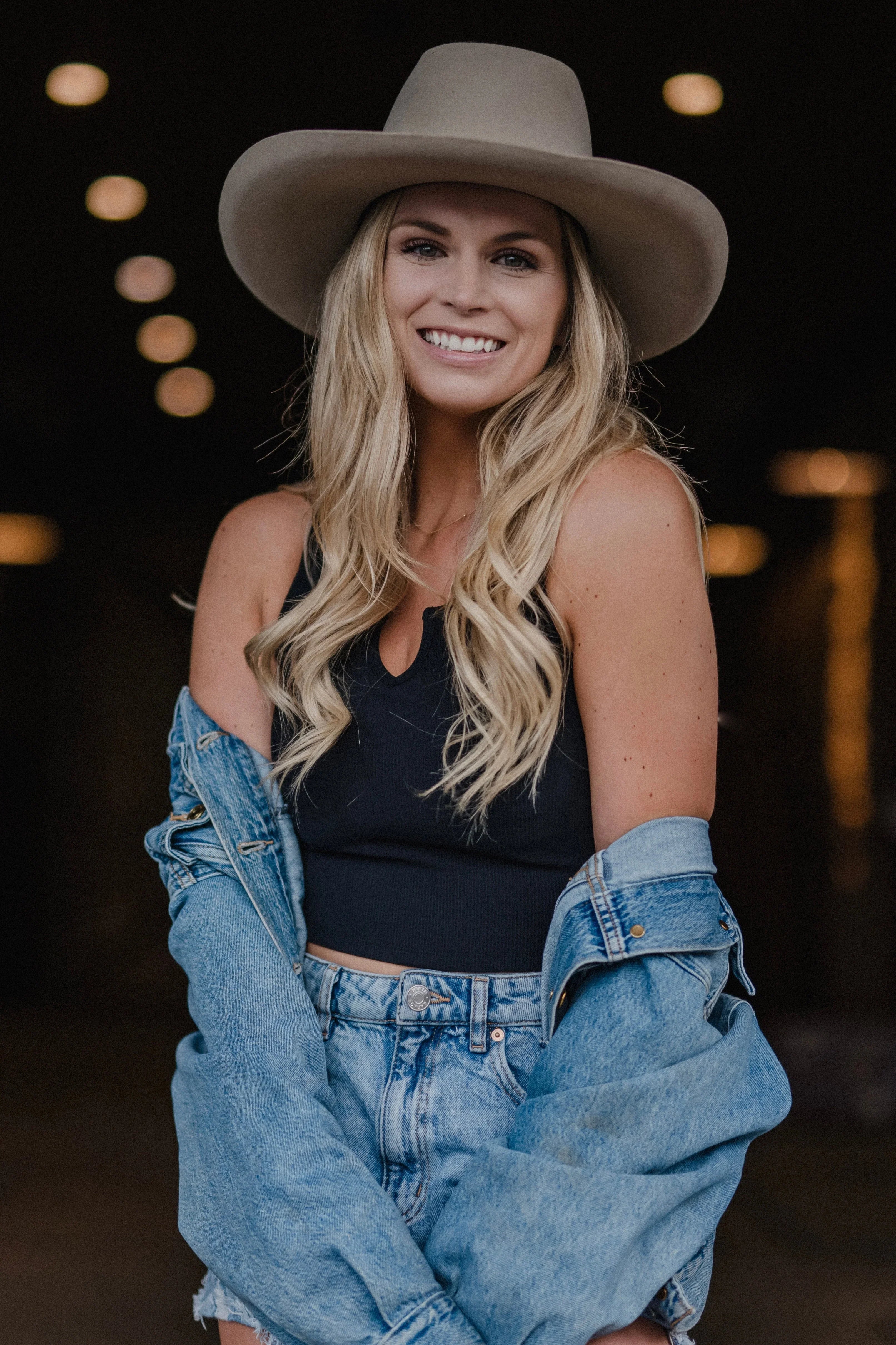 Smiling woman in wide-brim hat, black top, and denim jacket with long blonde hair