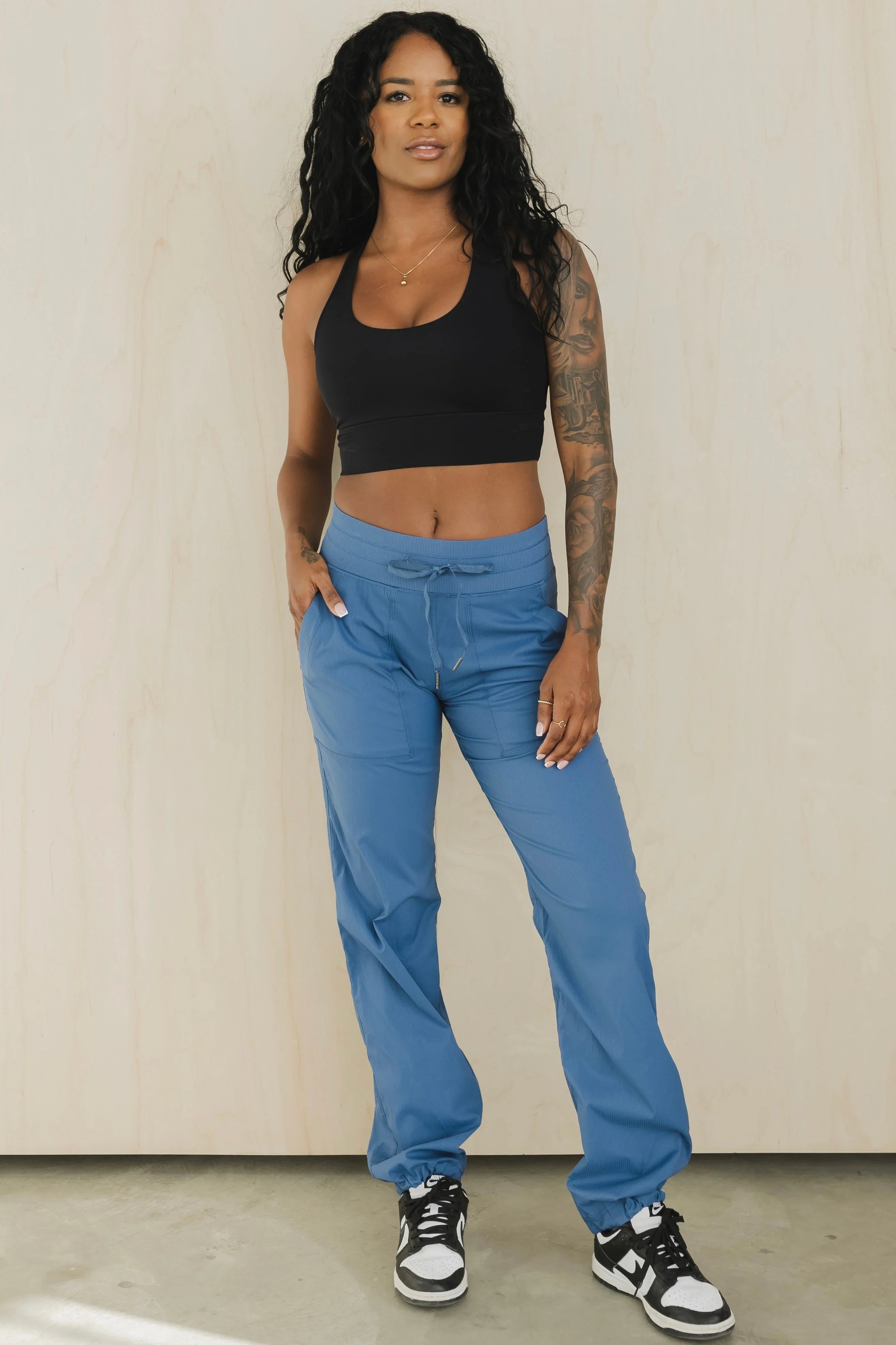 Woman in black crop top and blue jogger scrub pants standing against light wood wall