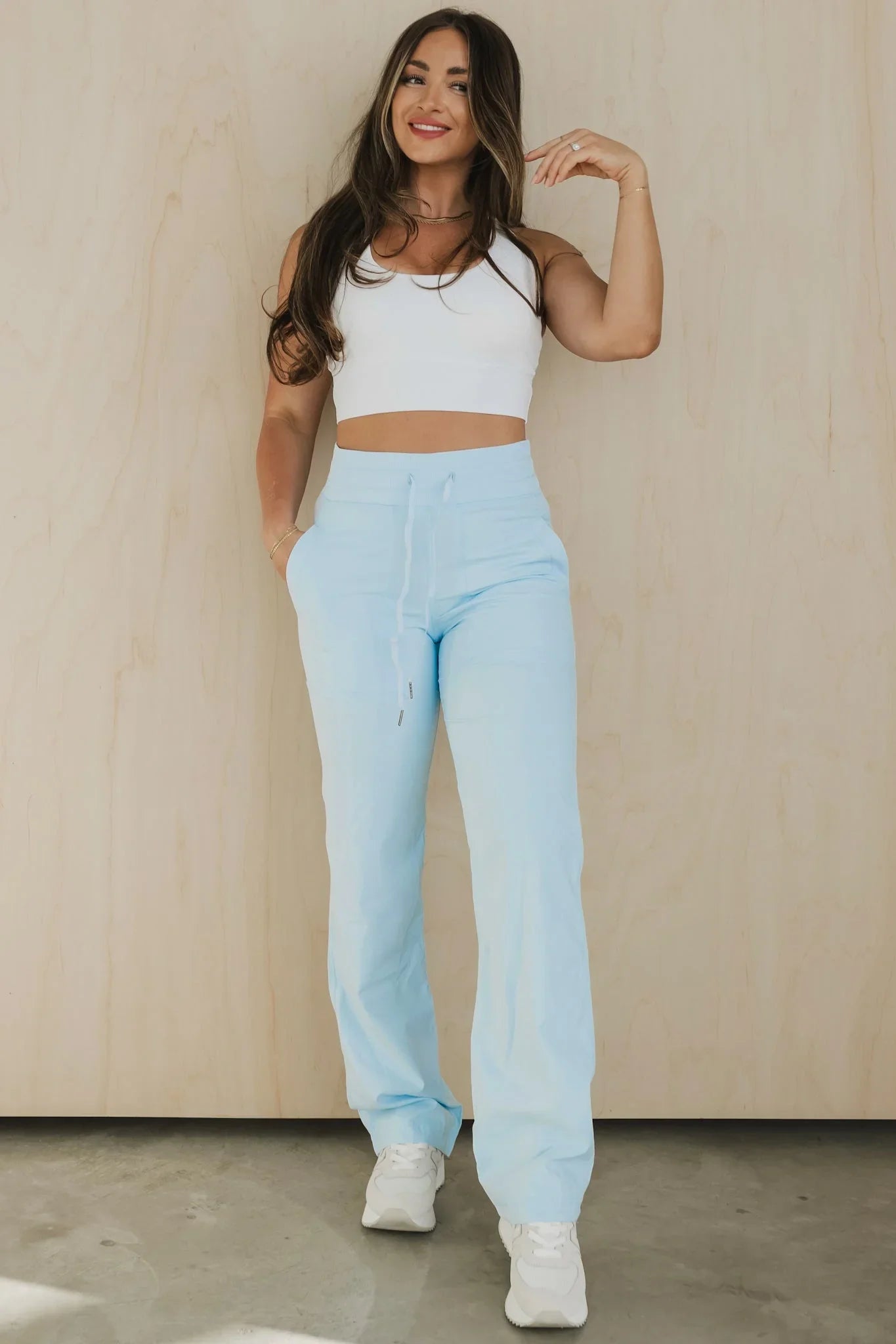 Woman in white crop top and light blue sweatpants poses against beige wall