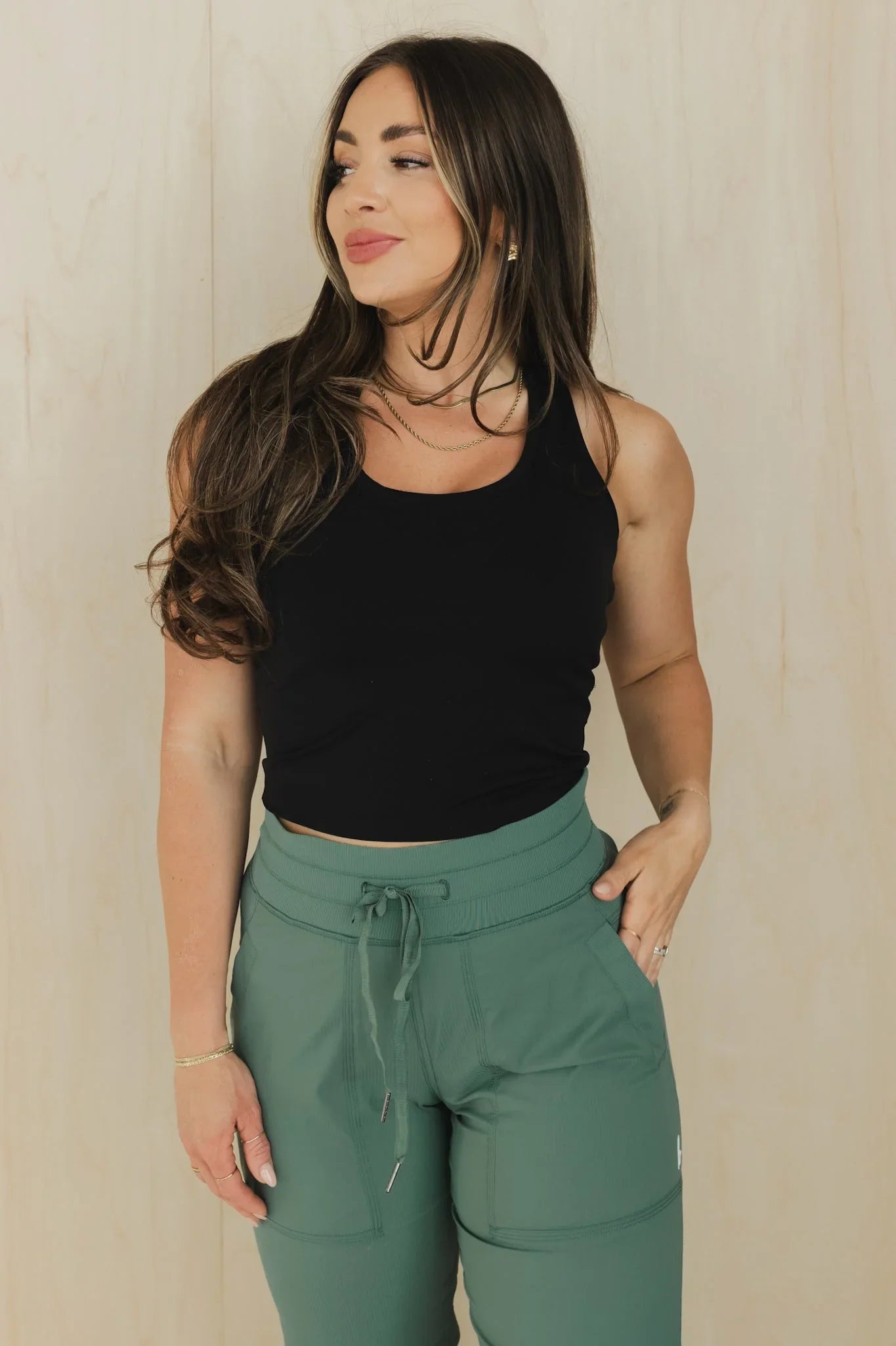 Woman in black tank top and green jogger pants standing against light wood background