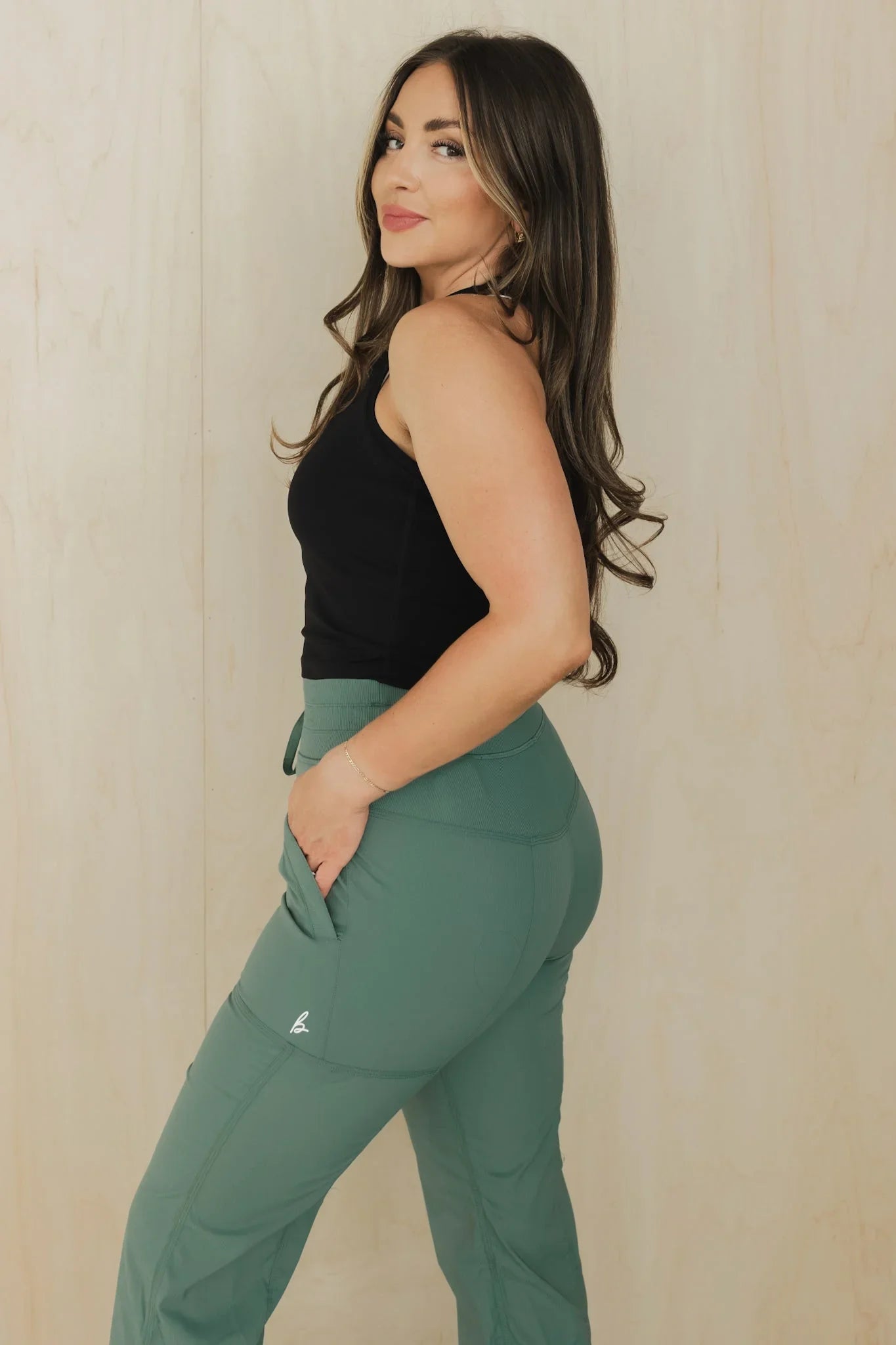 Woman in black tank top and green jogger pants posing against a light wood background