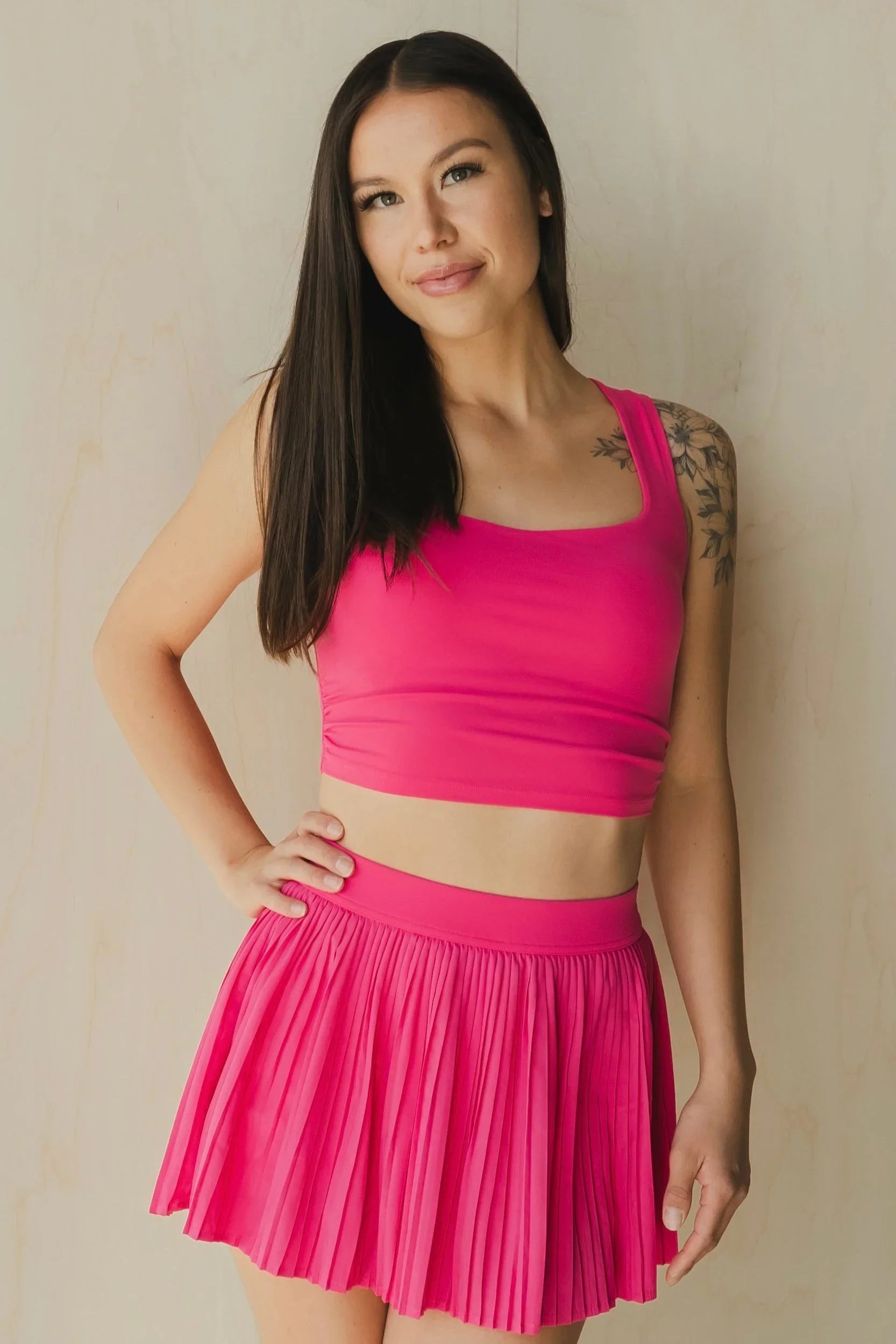 Woman wearing hot pink sports crop top and pleated skirt, standing against beige wall