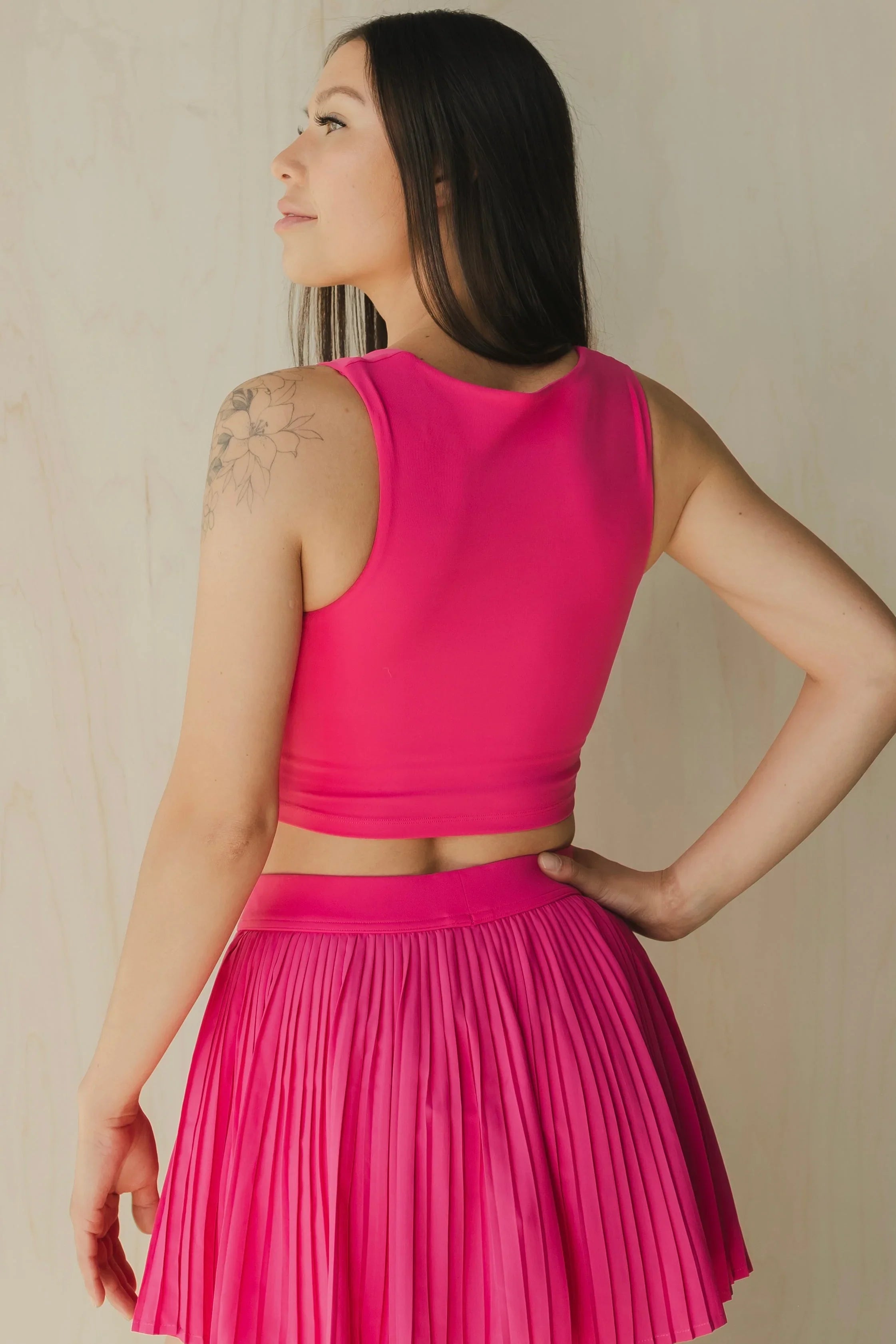 Woman in hot pink sleeveless crop top and pleated skirt, floral arm tattoo, neutral background