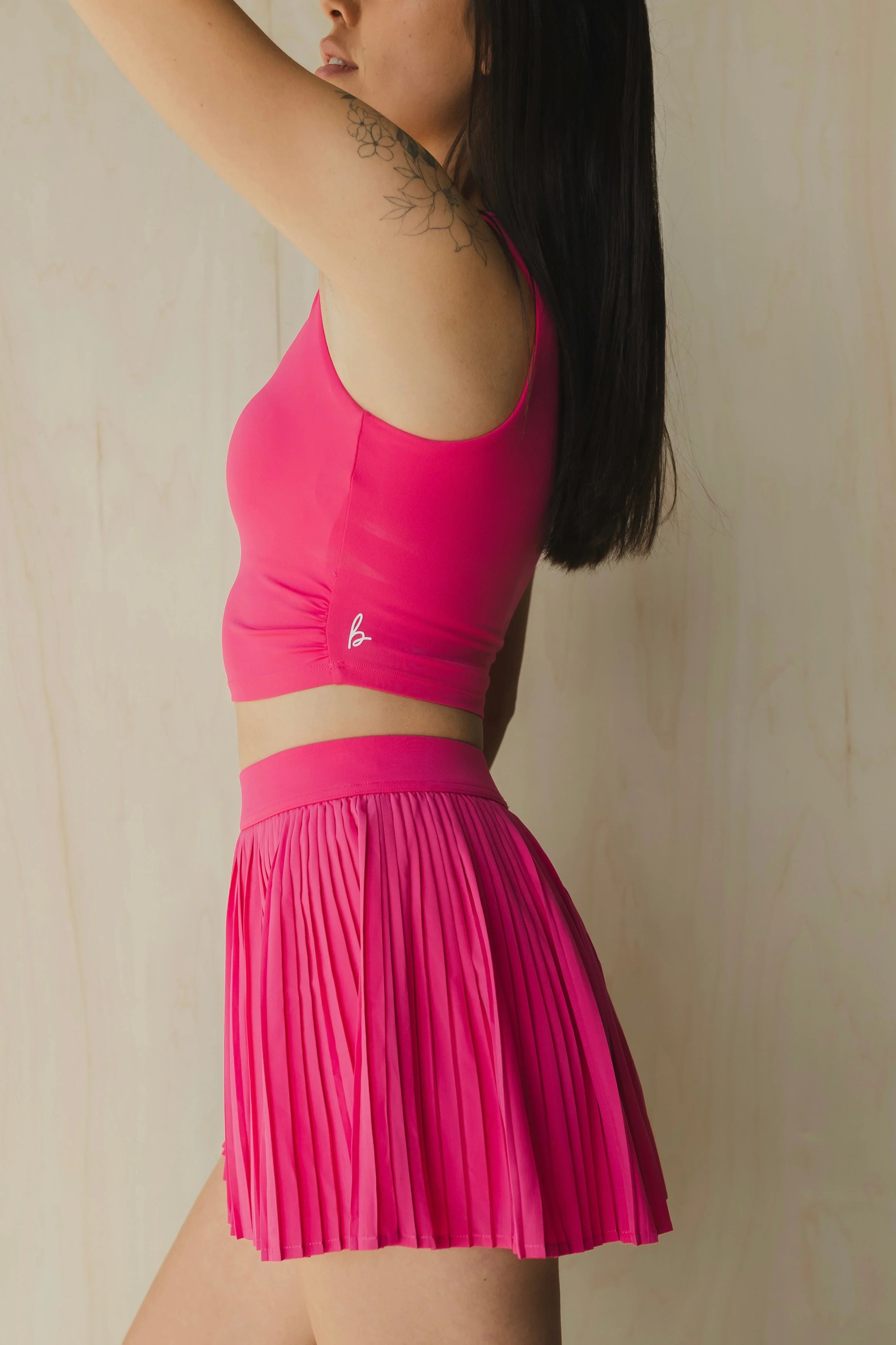 Woman in pink crop top and pleated mini skirt with floral tattoo, neutral background