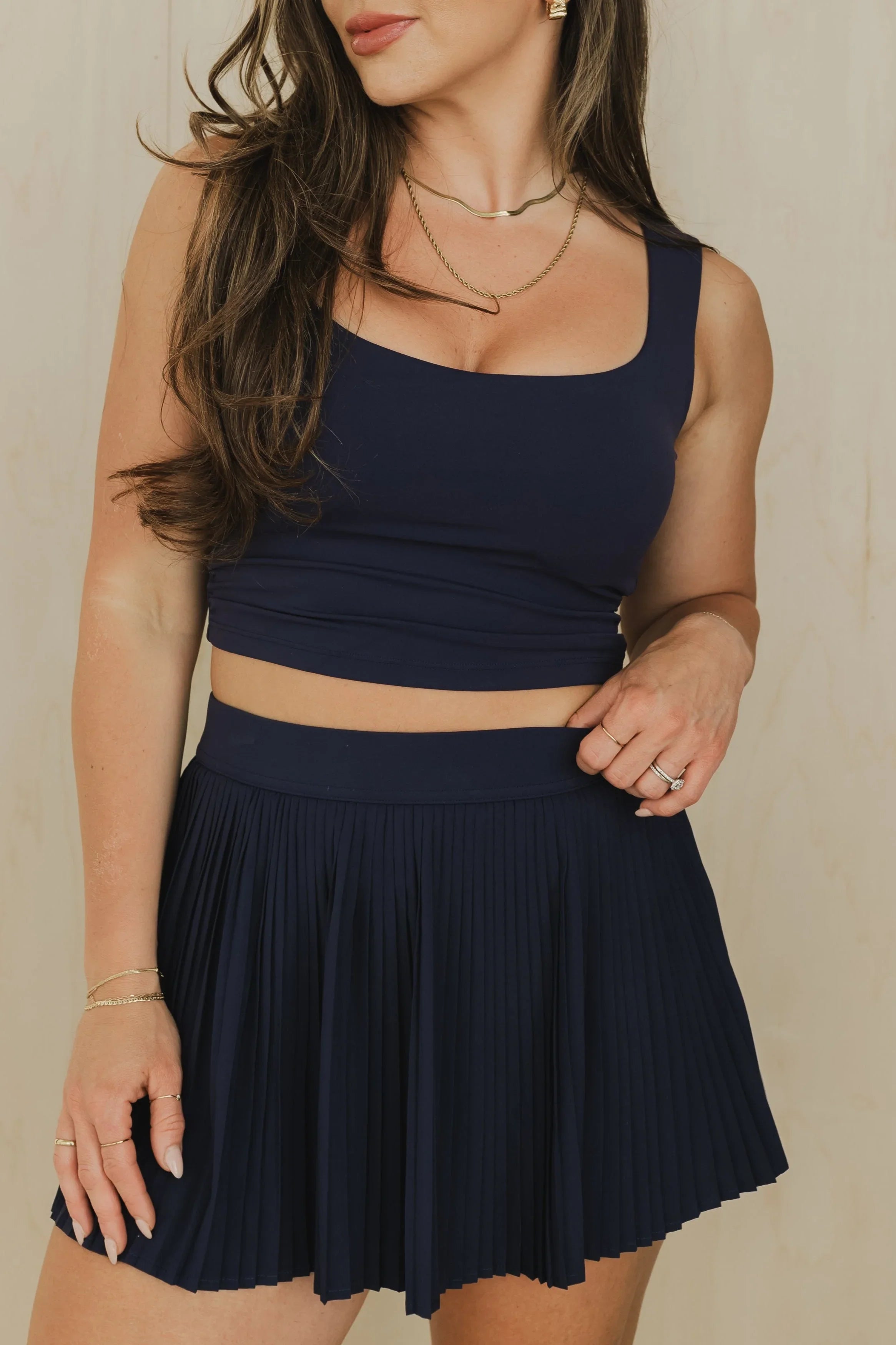 Woman wearing navy blue crop top and pleated skirt, gold jewelry, neutral background