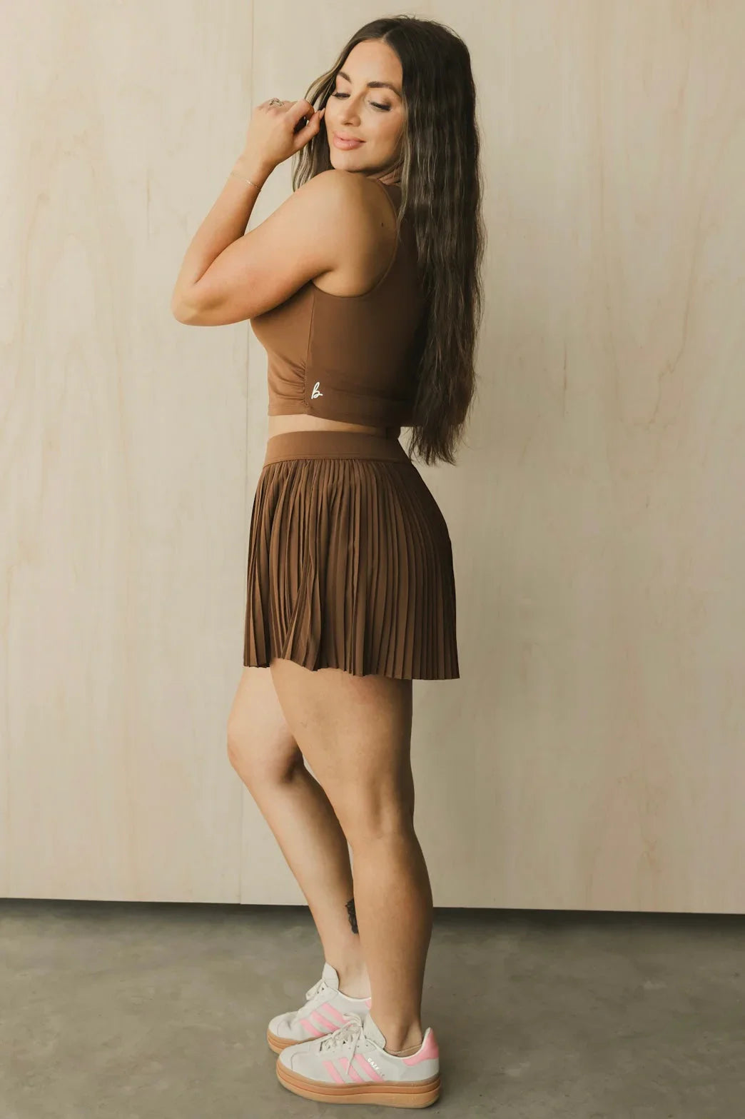 Woman in brown crop top and pleated skirt with long hair and white pink sneakers