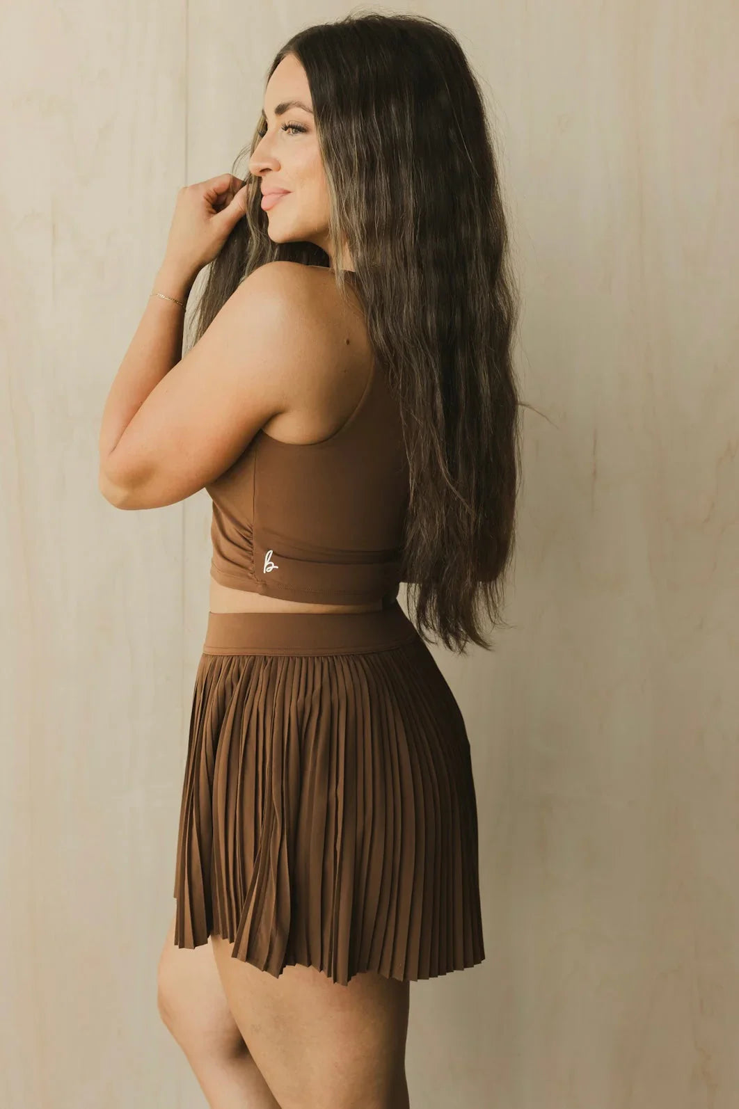 Woman in brown pleated skirt and crop top posing against beige background, athletic fashion